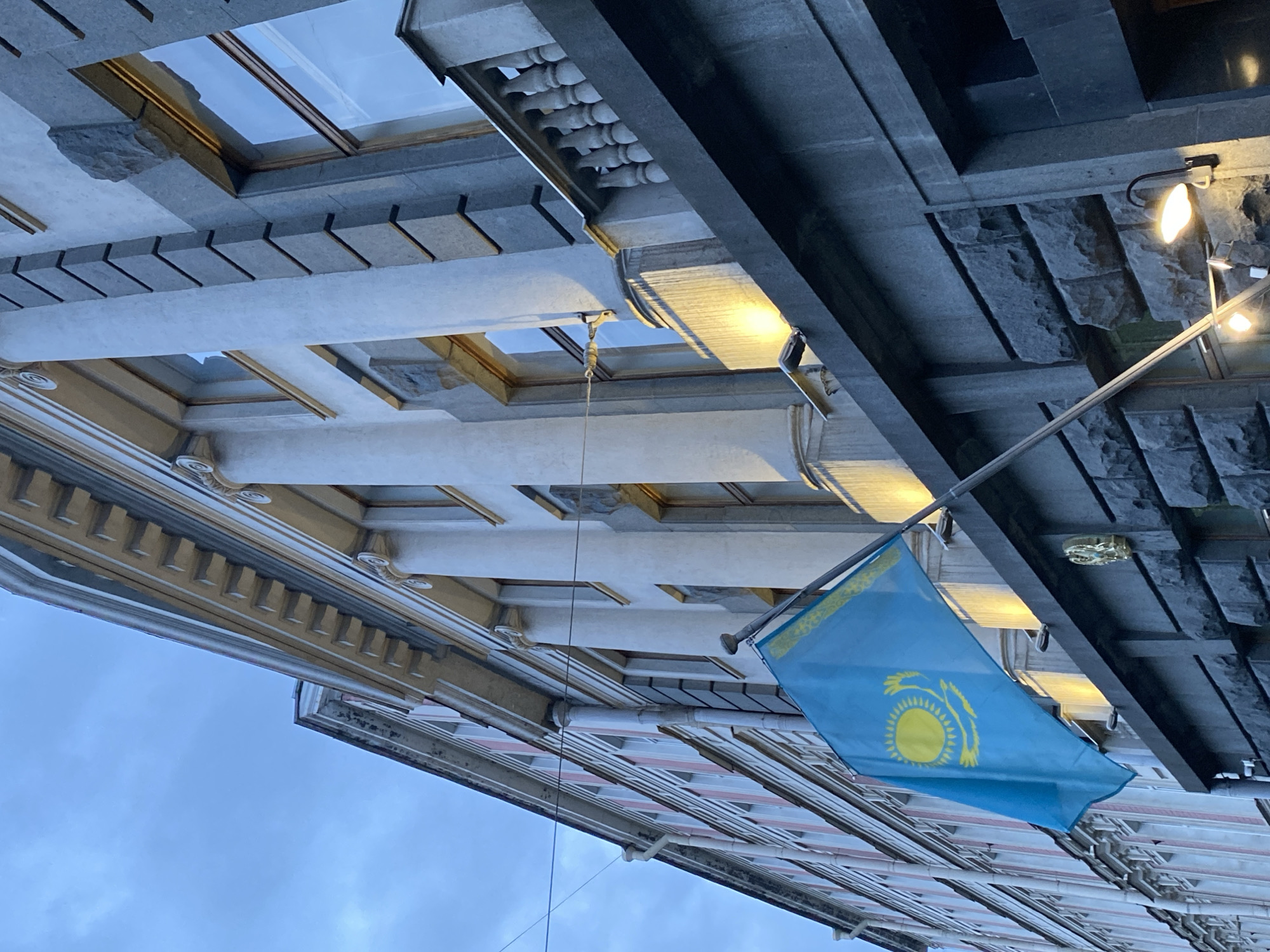 Here is a description of the image:
The image is a low-angle shot of buildings in Saint Petersburg, Russia. The sky is overcast, suggesting dusk or early morning. The main subject is the flag of Kazakhstan, which is prominently displayed. The buildings have decorative architectural features, including ornate cornices, moldings, and stone facades. Some of the buildings are illuminated by artificial lights. The architectural style is likely classical or neoclassical, typical of Saint Petersburg. The perspective is upward, emphasizing the height and grandeur of the buildings.