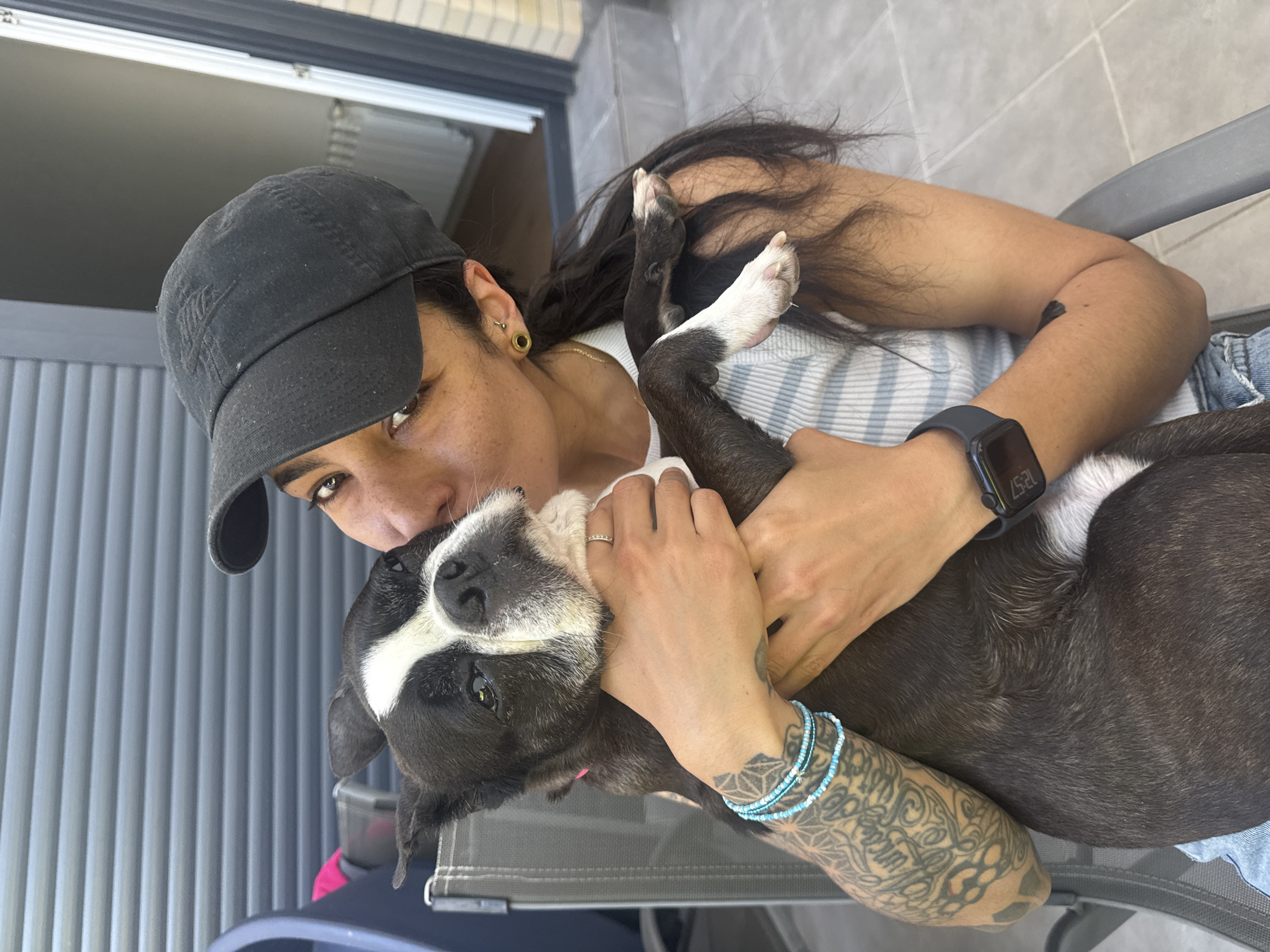 The image shows a person wearing a black cap, a white tank top, and blue jeans, embracing a black and white dog. The person has a gold earring, a watch on their left wrist, and blue beaded bracelets on their left forearm, which is also covered in tattoos. They are kissing the dog's head. The dog is mostly black with a white stripe on its face and white paws. The scene appears to be outdoors on a patio or deck with gray furniture and a sliding glass door in the background. The location is in the city of Unknown, Unknown.