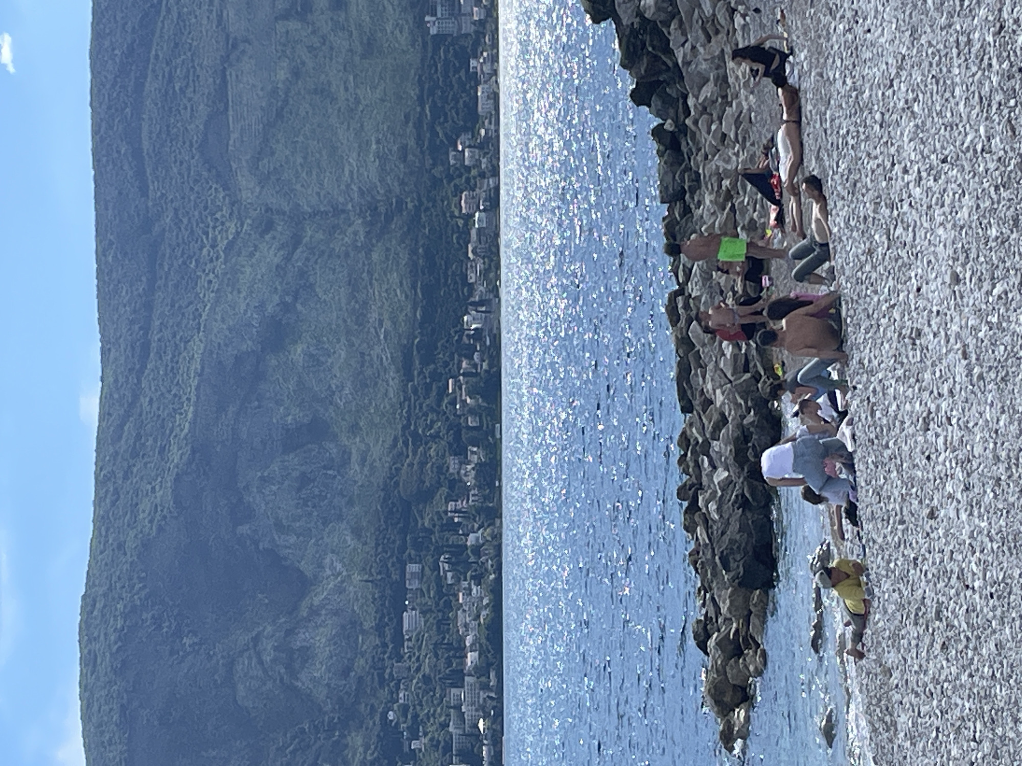 The image displays a vibrant coastal scene, likely in Herceg Novi, Montenegro, bathed in bright daylight. The primary focus is a pebbled or rocky beach in the foreground on the right, populated by a group of individuals.
On the rocky shore, several people are visible. Some are standing, others are sitting, appearing relaxed and engaged in leisure activities by the water's edge. One person in bright neon green shorts stands partially submerged in the sparkling blue water. Another, wearing a yellow top and light shorts, is bending down close to the water. The people are dressed in casual summer attire, including swimwear. The shoreline itself is composed of countless light-colored pebbles and larger, darker rocks forming a natural boundary with the sea.
The middle ground is dominated by a large body of water, likely the Adriatic Sea or a bay. Its surface gleams intensely with numerous bright reflections, indicating strong sunshine.
In the background, a dramatic landscape unfolds. A densely vegetated, towering mountain rises steeply, covered in a lush green canopy. At the base of this mountain, along the distant coastline, a town or settlement is visible, with buildings and structures nestled into the landscape. The sky above is a clear, brilliant blue, with a single small white cloud hovering near the top left corner. The overall impression is one of a sunny, pleasant day at a scenic coastal location. There is no visible text in the image.