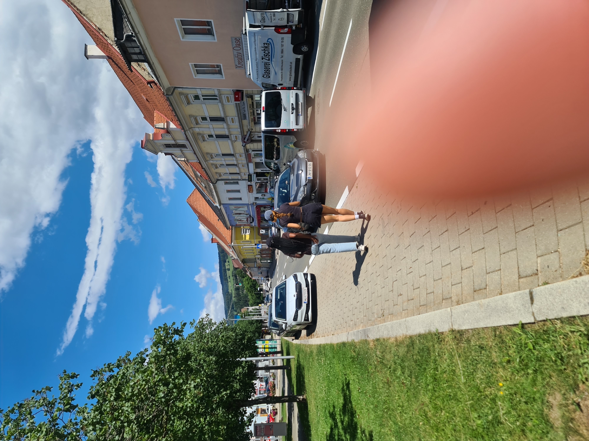 A medium shot captures a street scene in Sankt Marein bei Neumarkt, Austria, on a clear day with scattered clouds. Two young women walk away from the camera along a brick sidewalk. The woman in front wears a black t-shirt and black shorts, with her dark hair in a braid. The woman behind her wears a black t-shirt and light blue jeans, with her dark hair in braids. They are walking on a sidewalk bordered by a grassy area on the left. Across the street, a row of buildings with colorful facades line the road. Several vehicles are present on the street, including a white Skoda, a dark gray hatchback, two vans, and a truck. A white van with "Glaserei Zischka" written on it is parked on the right side of the street. In the background, a green hill with scattered trees and houses is visible under a bright blue sky. The overall impression is a sunny day in a European town.