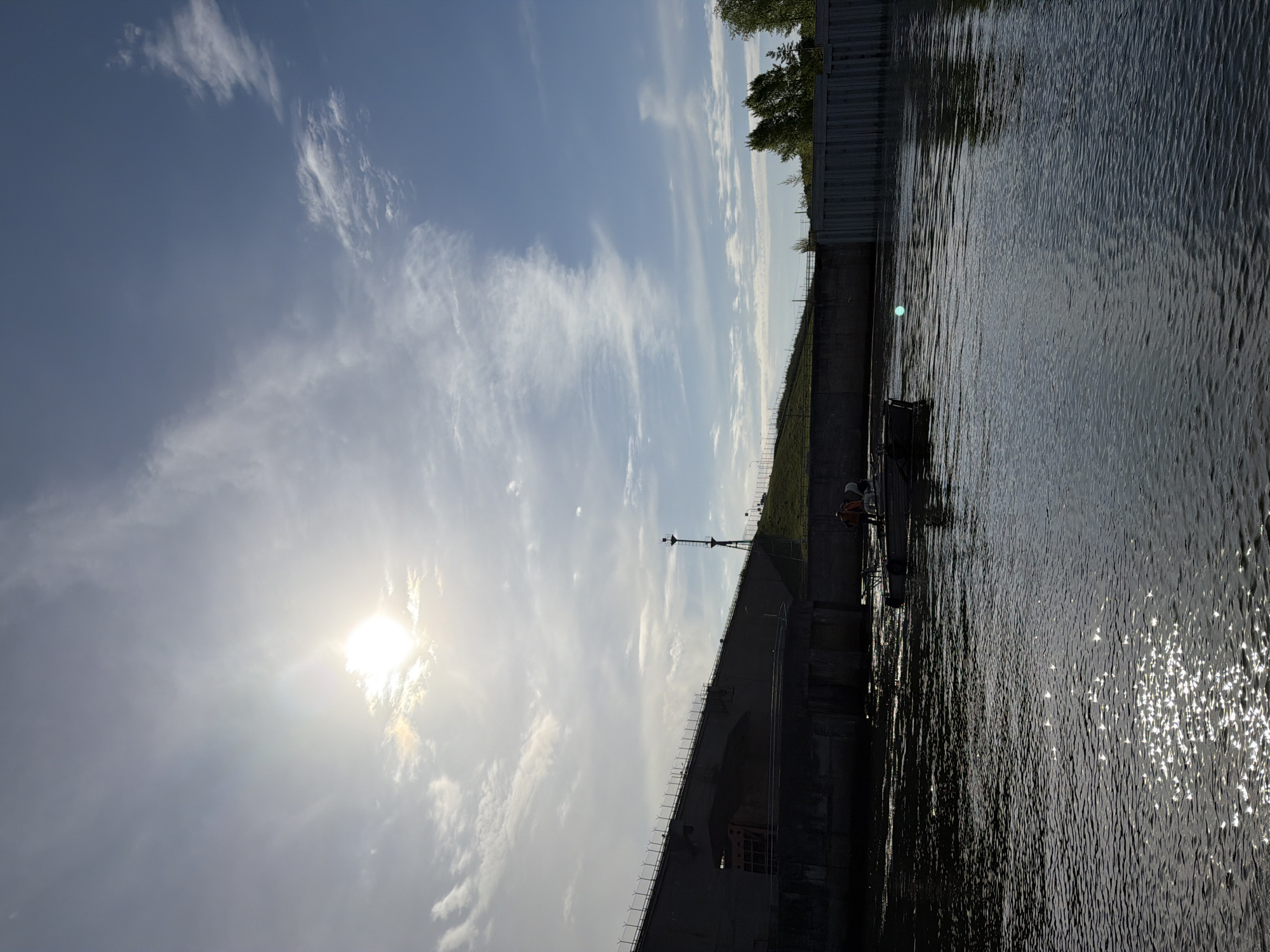 A photograph taken in Chaikovsky, Russia, shows a body of water next to a concrete structure. The sun is visible through the clouds in the sky. A transmission tower is atop the concrete structure. Two workers are on the platform.