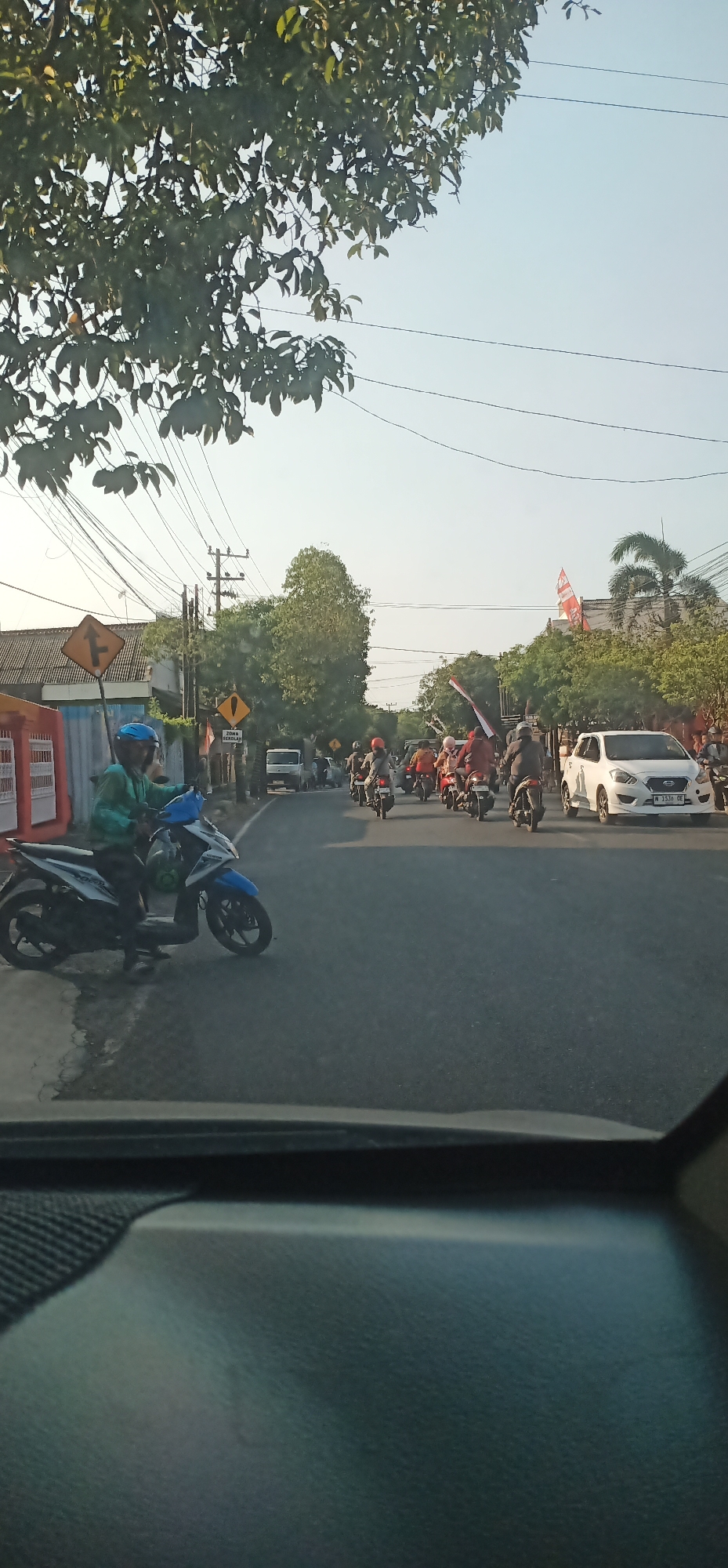 The image captures a street scene in Probolinggo, Indonesia, on a bright, clear day. In the foreground, the dashboard of a vehicle is visible, suggesting the photo was taken from inside a car. The street is moderately busy with a convoy of motorcycles, some carrying passengers, heading down the road. Several of the motorcycles appear to be decorated with Indonesian flags. On the left side of the street, a motorcycle rider wearing a green jacket and blue helmet is stopped. Further down the road, there is a white car and a small truck. Trees line the sides of the street, and overhead power lines are visible. Signs, including a yellow directional sign and a sign indicating "ZONA SEKOLAH" (School Zone), are present along the roadside. Buildings with red and white facades are also visible in the background. The overall impression is a procession or gathering of motorcycles on a typical Indonesian street.