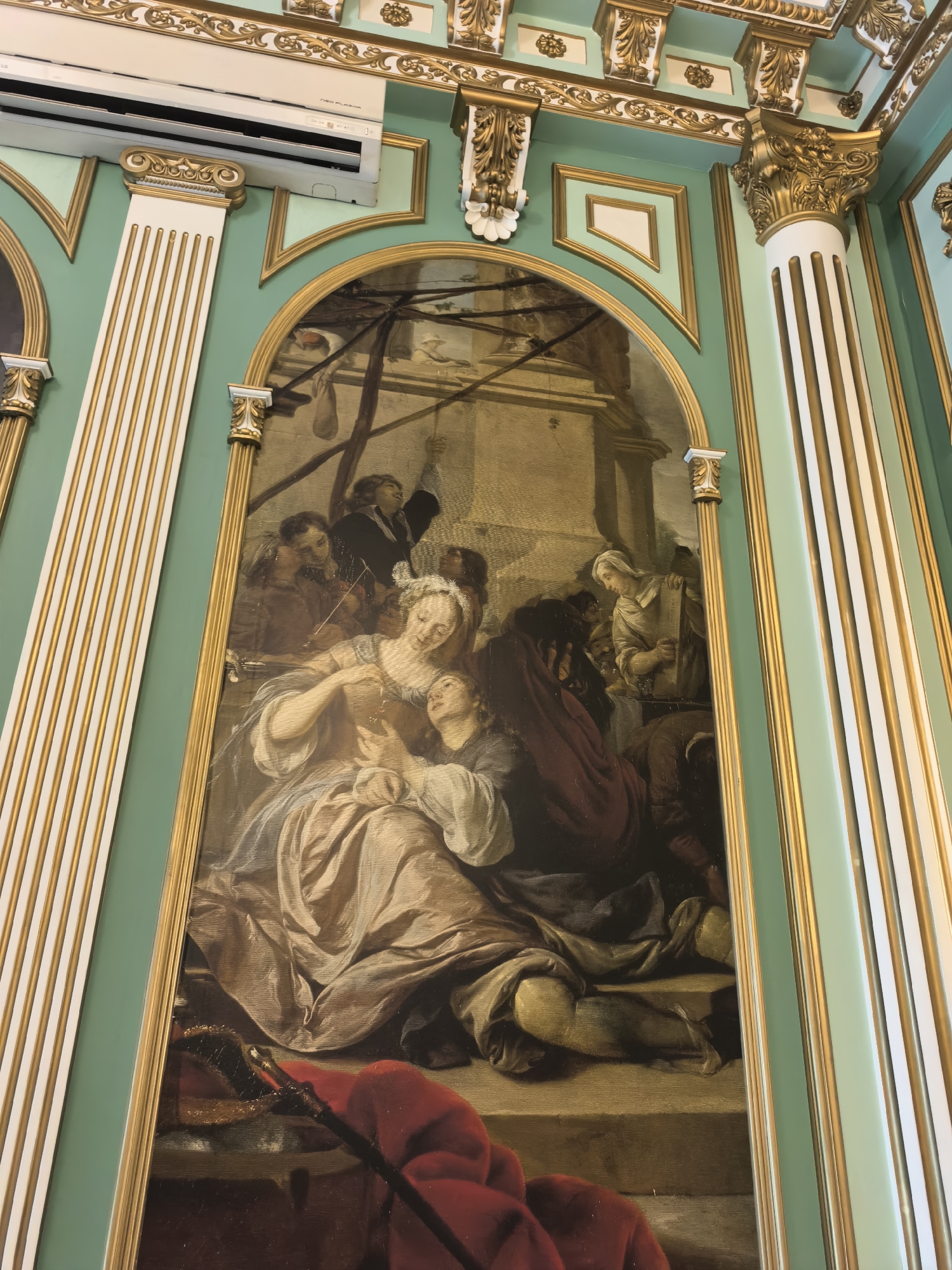 An indoor, eye-level, medium shot captures a painting depicting a religious or historical scene. The painting is framed by ornate green and gold molding and is set against a pale green wall with decorative gold trim. A white and gold column is visible on the left side, and another on the right. The painting itself shows a group of people, mostly women, gathered around a central figure who appears to be holding a child. The central figure is wearing a light-colored dress and her head is tilted back. A large red cloth is draped in the foreground. An air conditioning unit is mounted high on the wall in the upper left corner of the image. The lighting is bright and even, suggesting artificial illumination within a building.
