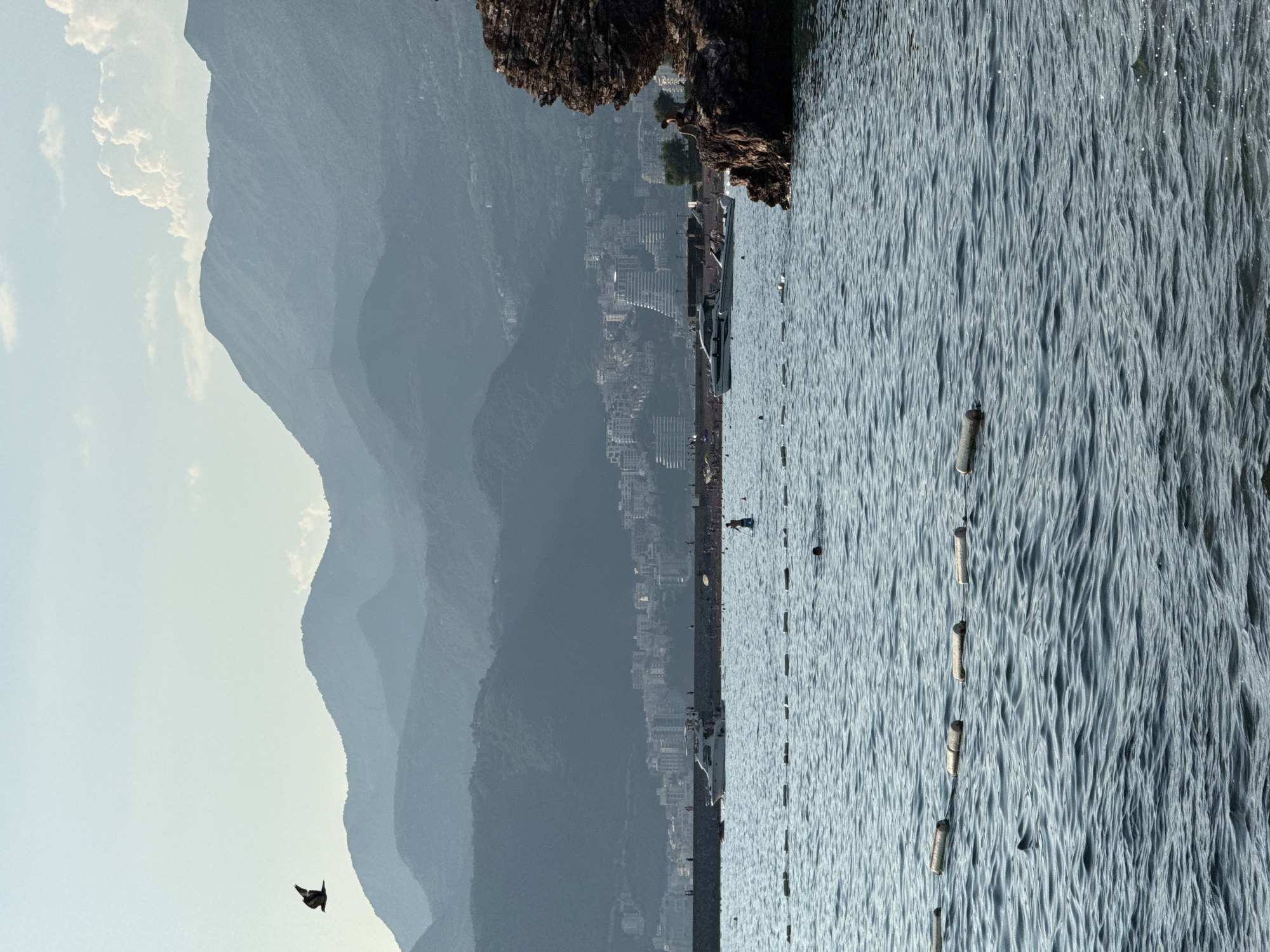 The image depicts a coastal scene in Kamenovo, Montenegro. In the foreground, a choppy, blue sea stretches out, marked by a line of buoys. The water's surface is textured with small waves, creating a shimmering effect. A small, dark object, possibly a bird in flight, is visible in the lower left corner of the frame against a pale sky.
In the mid-ground, a rocky outcrop juts into the water on the right. Beyond this, a town with many buildings is nestled against a backdrop of hazy, layered mountains. A white boat is docked near the shore, and several figures are present on the waterfront, suggesting a mild, daytime atmosphere. The overall impression is of a serene, possibly warm day at a coastal resort town. There is no visible text in the image.
