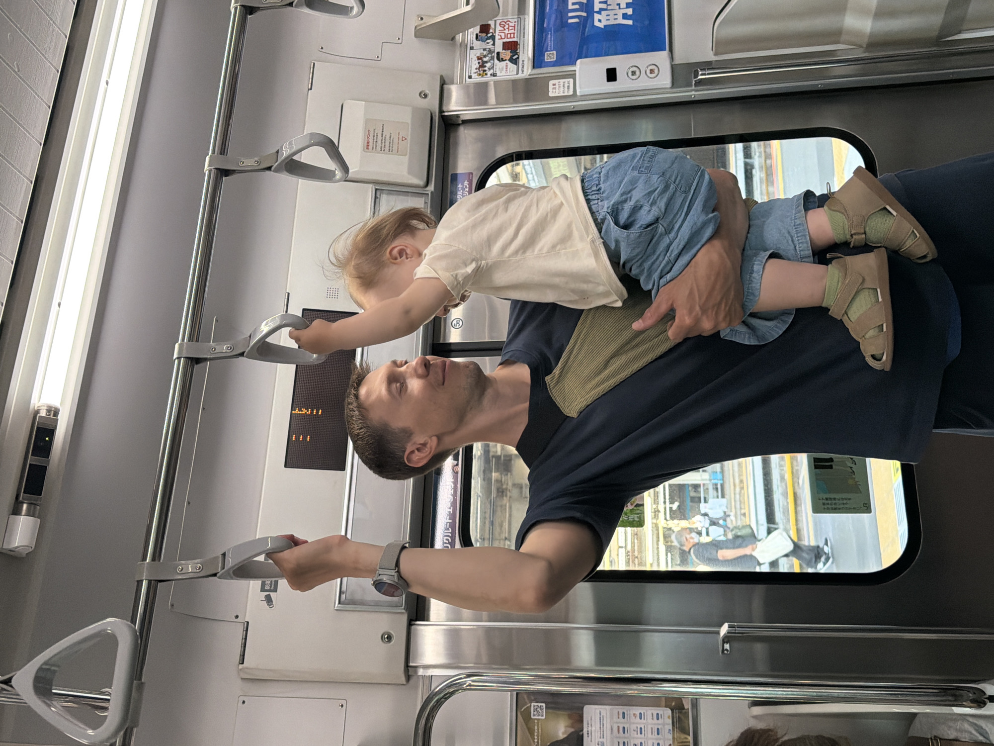 A man is holding a toddler on a train in Yokohama, Japan. The man, wearing a dark blue t-shirt and a light green strap across his chest, is looking to his right with a slight smile. He has a watch on his left wrist. The toddler, wearing a light-colored t-shirt and denim shorts, is facing away from the camera, reaching up towards a handrail. The toddler is wearing light brown sandals and green socks. They are on a train with white walls and metal poles. Outside the train window, a person wearing a face mask and carrying a white bag is walking on a platform. Various signs with Japanese text are visible on the train walls and outside the window.