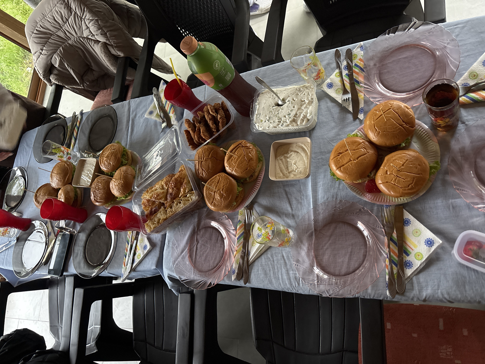 A table laden with food is the focal point of this image. Burgers, fried chicken, salad, and pastries are arranged amidst plates, cups, cutlery, and a large bottle of juice. The table is covered with a light blue, striped tablecloth. The setting is likely indoors, given the presence of chairs around the table and a partial view of a window showing greenery outside. The location is not discernible as Zlatograd, Bulgaria, from the image. No people are visible in the frame.