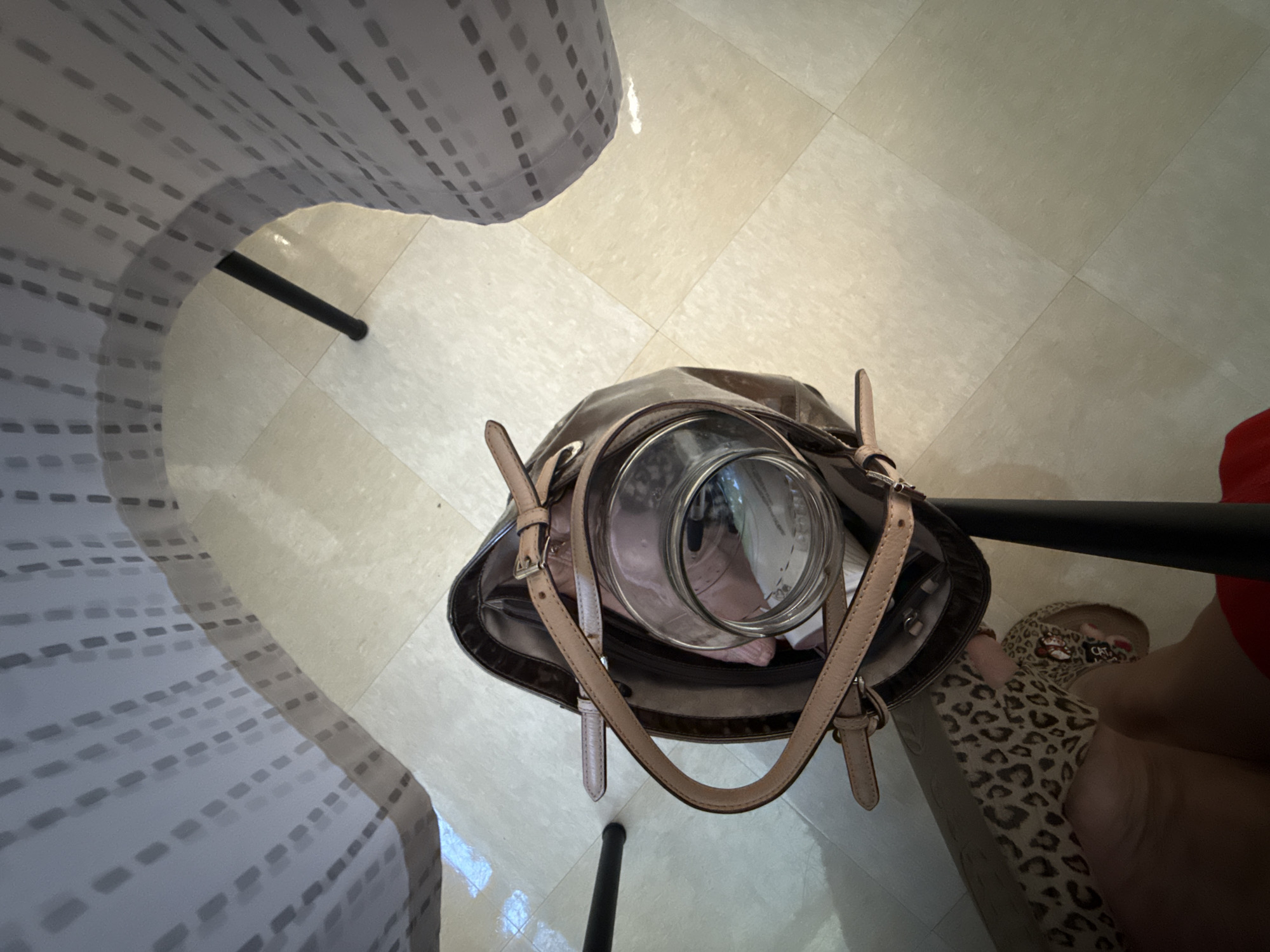 A high-angle, eye-level shot shows a brown leather tote bag resting on a light-colored tiled floor. The bag contains a clear glass jar and some light-colored items, possibly clothing. A person's legs and feet are visible in the lower left of the frame, one leg clad in a leopard-print material and the other in a red garment. The person is wearing a sandal adorned with cartoon characters. The scene appears to be indoors, possibly under a table or desk, as a patterned, sheer curtain is draped from above, casting shadows. The floor tiles are arranged in a grid pattern. The lighting is dim, with some areas brighter than others due to artificial light sources. There are no identifiable landmarks or features to indicate a specific location in Rome, United States of America.