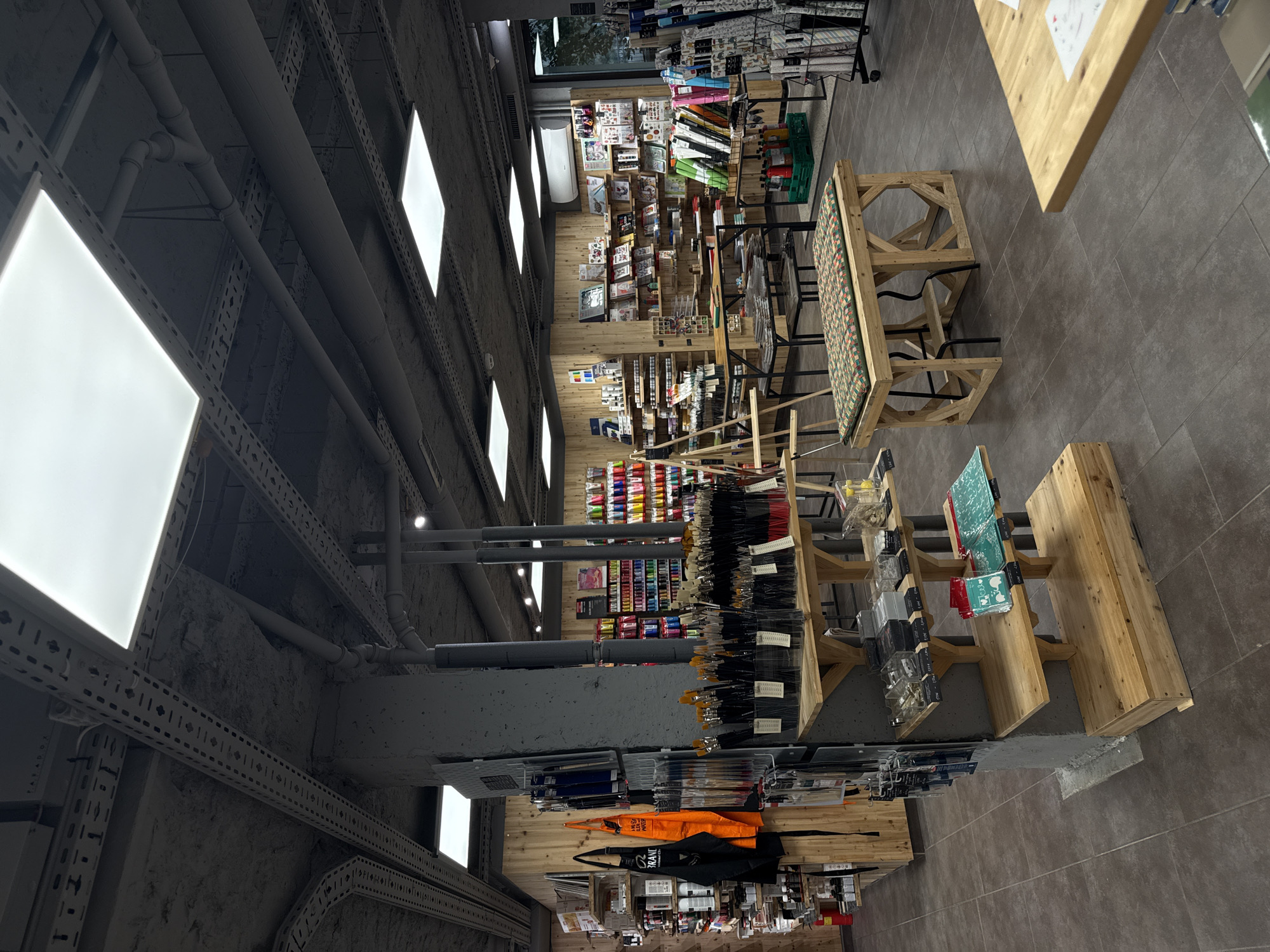 This image captures the interior of what appears to be an art or craft supply store in Sofia, Bulgaria, characterized by an industrial-chic aesthetic. The perspective looks down an aisle, showcasing various merchandise displays.
The ceiling features exposed concrete, grey pipes, and white rectangular LED panel lights that provide bright illumination. Metal grates and perforated channels for cables or utilities are also visible along the ceiling. The walls are a textured, rough concrete finish, while the floor is laid with dark grey tiles.
Numerous wooden display shelves and racks fill the store, holding a wide assortment of products. On the left side, shelves are stocked with various paint tubes and bottles in a spectrum of colors. Further along, a large collection of paintbrushes with black handles is neatly displayed. Other shelves contain items like rolls of decorative paper, sketchbooks, drawing pads, stencils, rulers, and various small craft accessories. An orange tote bag and a black apron or bag, partially displaying the text "ERRANT" (possibly "FERRANT" or a similar brand name), hang on a display near the bottom.
In the middle of the store, a long wooden bench or table with a multi-patterned cushion provides a seating area or additional display space. A plain wooden table is partially visible in the right foreground. No people are present, indicating a quiet moment in the store. The overall impression is an organized and well-stocked environment for artistic and craft endeavors.