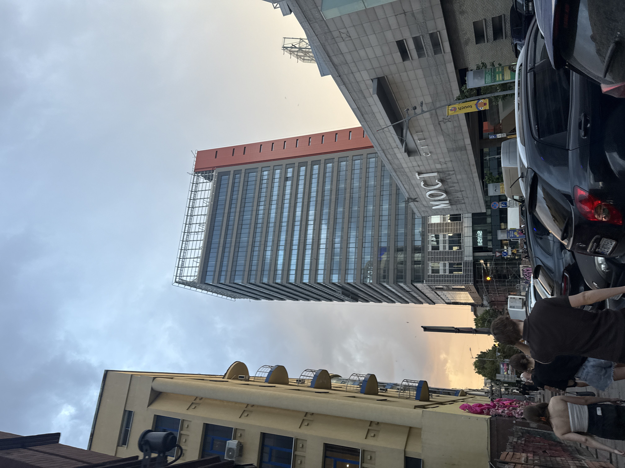 The image depicts a street scene in Dnipro, Ukraine, likely near the "Most City" complex. A tall, modern glass-fronted skyscraper with a red accent panel dominates the center of the frame. To its left, a yellow building with rounded balconies is visible. The foreground is filled with parked cars, and a group of people are walking away from the camera on a sidewalk. The sky is overcast with hints of sunset colors. The overall impression is an urban environment captured during the evening.