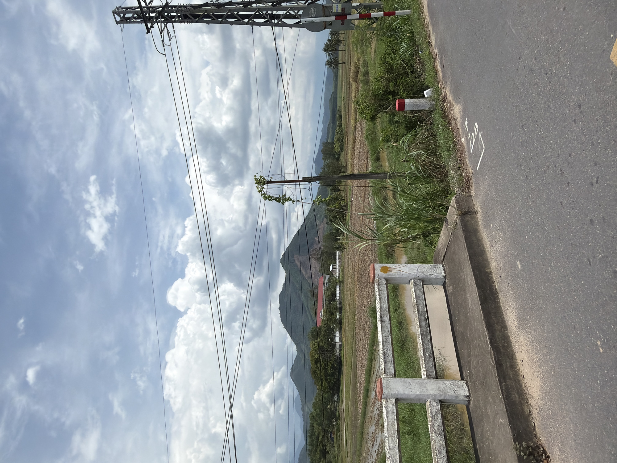 The image shows a rural landscape with a road in the foreground. A white concrete bridge railing with red and white posts runs along the left side of the road. Beyond the railing, there is a field of dry crops or harvested rice paddies with some water in them. In the middle ground, there are trees and a building with a red roof. Further in the background, rolling green hills and mountains are visible under a cloudy sky. A power line tower and wires are prominent in the upper right of the image. The road has white markings on it, including what appears to be a number "428". The scene is likely located in or near Diên Thọ, Vietnam, given the rural scenery and architecture.