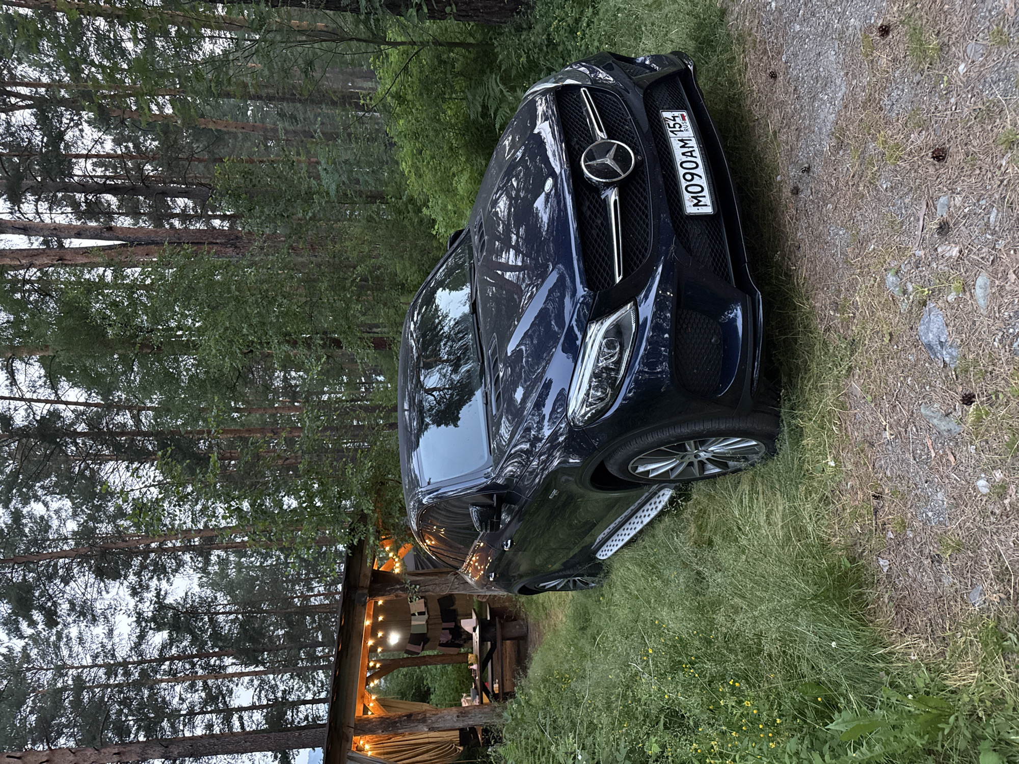 A dark blue Mercedes-Benz GLE SUV is parked on a gravel and grass area next to a wooden gazebo in a forest. The gazebo is illuminated with string lights and has a table and benches inside. The car's license plate reads "M090AM 154 RUS". The surrounding environment is a dense forest with tall pine trees and lush green undergrowth. The ground is covered with pine needles, dry leaves, and scattered rocks. The scene appears to be a serene outdoor setting, possibly a campsite or recreational area in Cheposh, Russia. There are no people visible in the image.