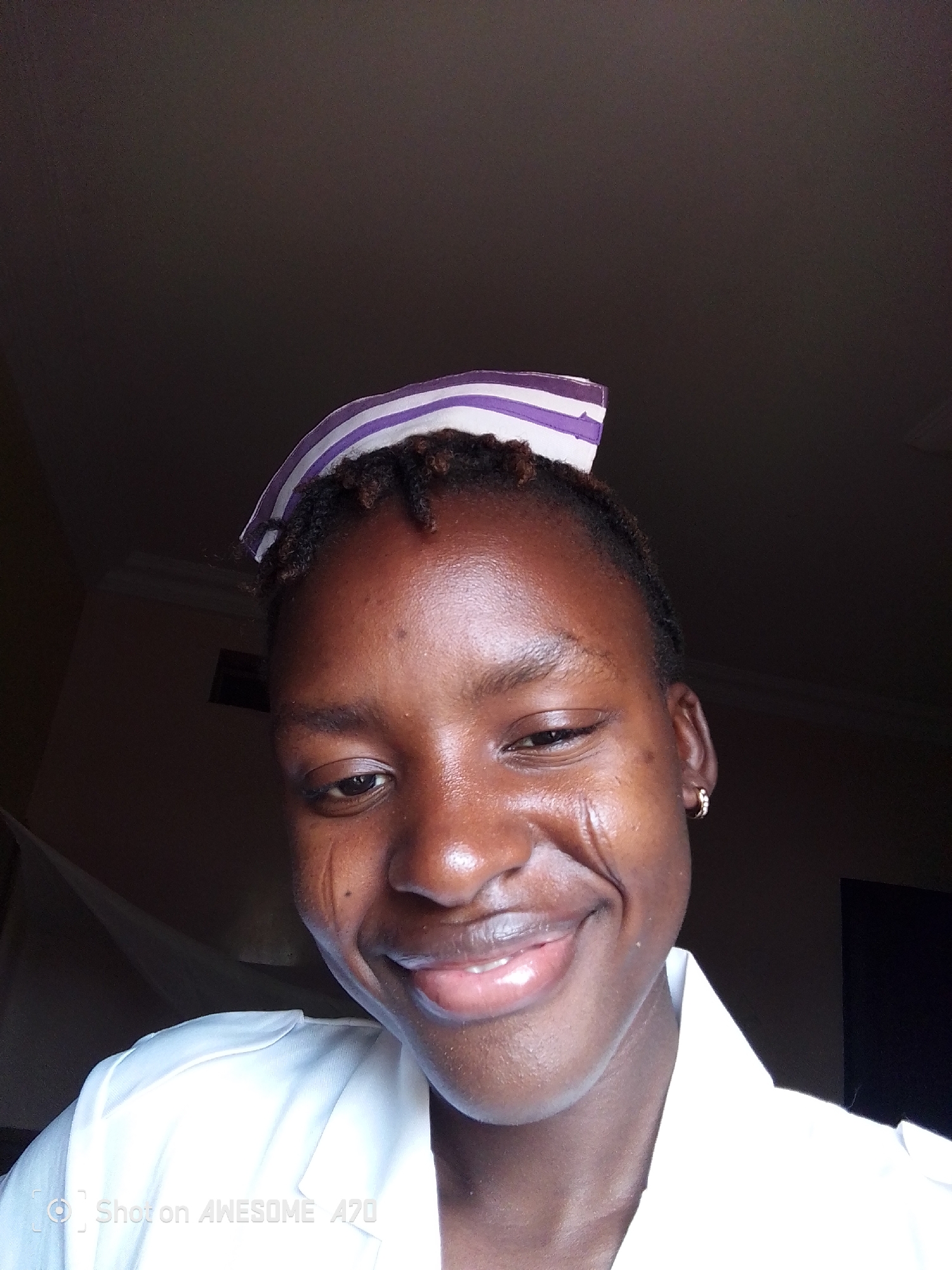 A close-up shot depicts a young woman, potentially in her early twenties, wearing a white uniform and a striped head covering that suggests a nursing or medical profession. The woman has dark skin, short dark hair visible under the cap, and a single earring in her left ear. She is smiling. There are visible marks or lines on her left cheek. The background is dark and indistinct. The city of Unknown, Unknown, cannot be determined from this image.