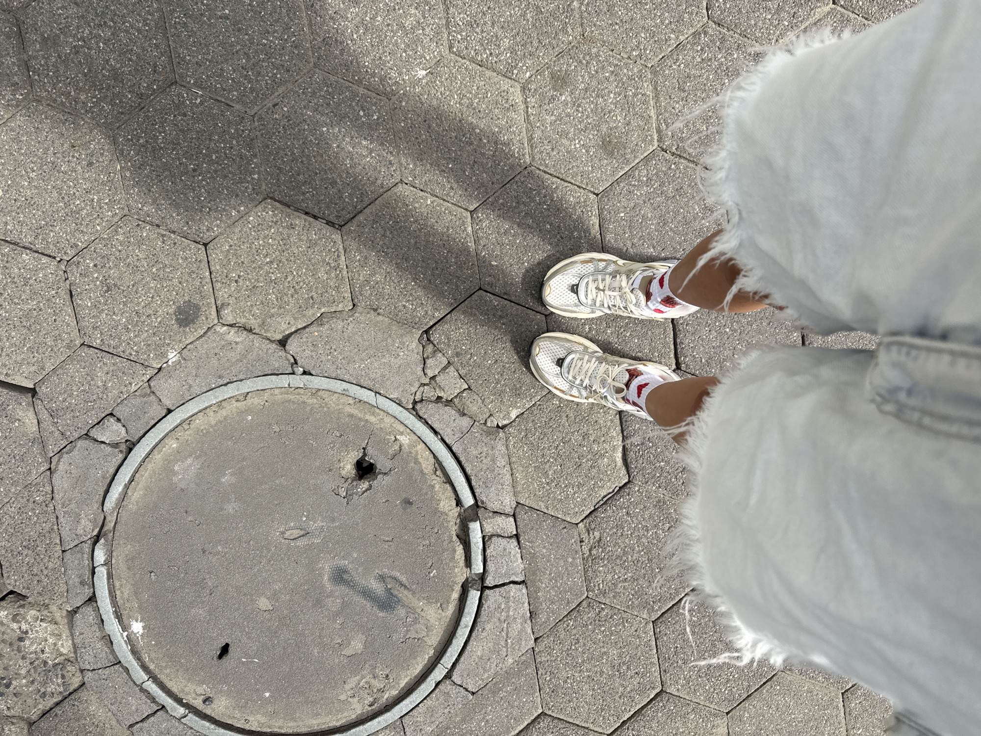 This is an overhead shot of a person's legs and feet standing on a hexagonal paver walkway, with a manhole cover in the lower left quadrant. The person is wearing white sneakers with red accents and light-colored, frayed denim shorts or a skirt. The walkway is made of dark gray hexagonal pavers, some of which are cracked and uneven. The manhole cover is round, made of gray concrete with a metal rim, and shows signs of wear, including small holes and scuff marks. Sunlight casts shadows from the pavers and what appears to be an unseen object, suggesting it is daytime. The scene is likely outdoors in an urban or park setting. There is no visible text in the image.