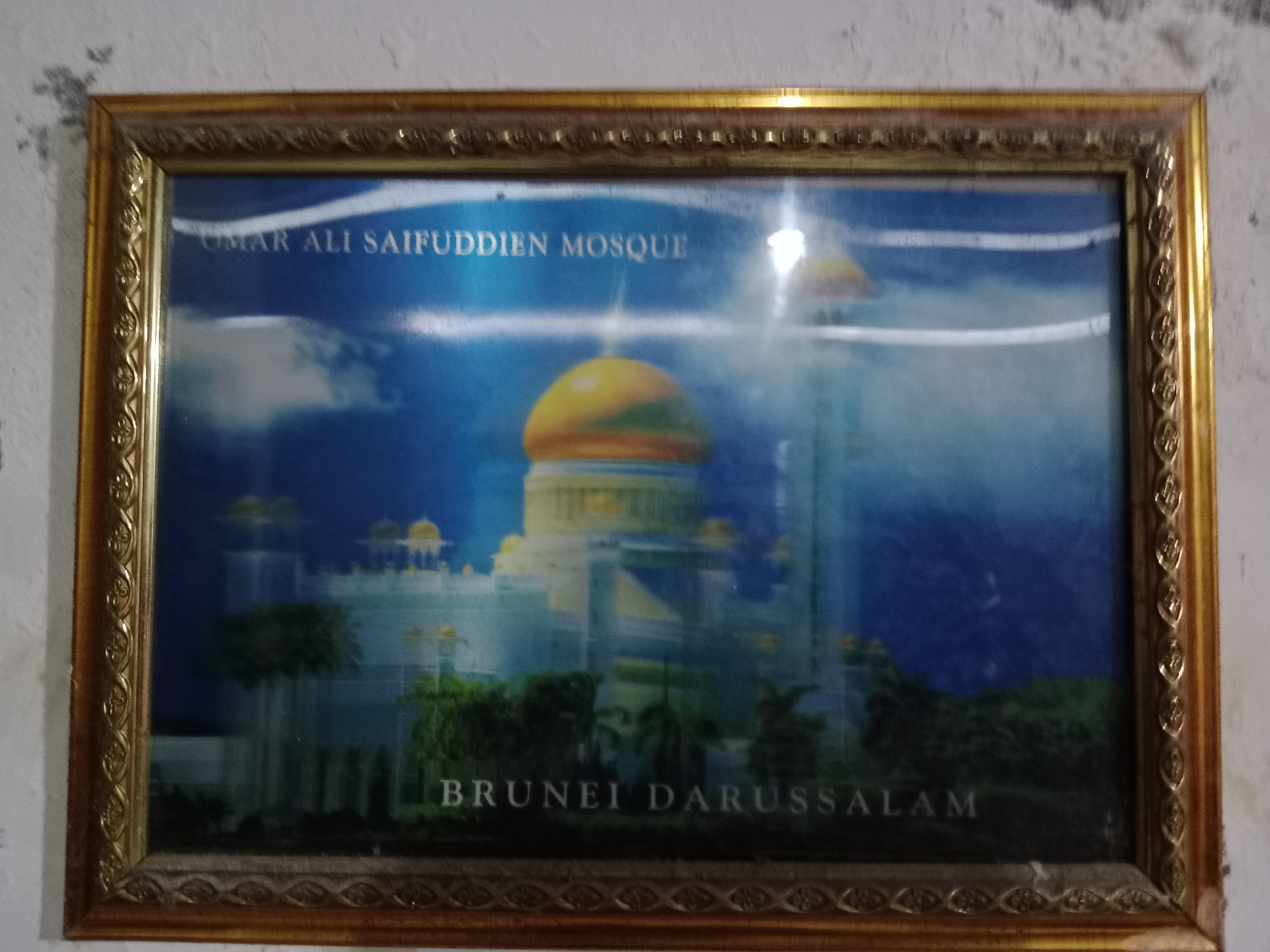 The image shows a framed picture on a wall. The picture is of the Omar Ali Saifuddien Mosque in Brunei Darussalam. The mosque is depicted with its iconic golden dome and minarets, surrounded by water and greenery. The sky is a vibrant blue with some clouds. The text "OMAR ALI SAIFUDDIEN MOSQUE" is visible at the top of the image, and "BRUNEI DARUSSALAM" is visible at the bottom. The frame is ornate and gold-colored. The wall behind the picture appears to be textured and slightly discolored. The location of the image is stated to be in the city of Ringinrejo, Indonesia, though the picture itself depicts a location in Brunei Darussalam.