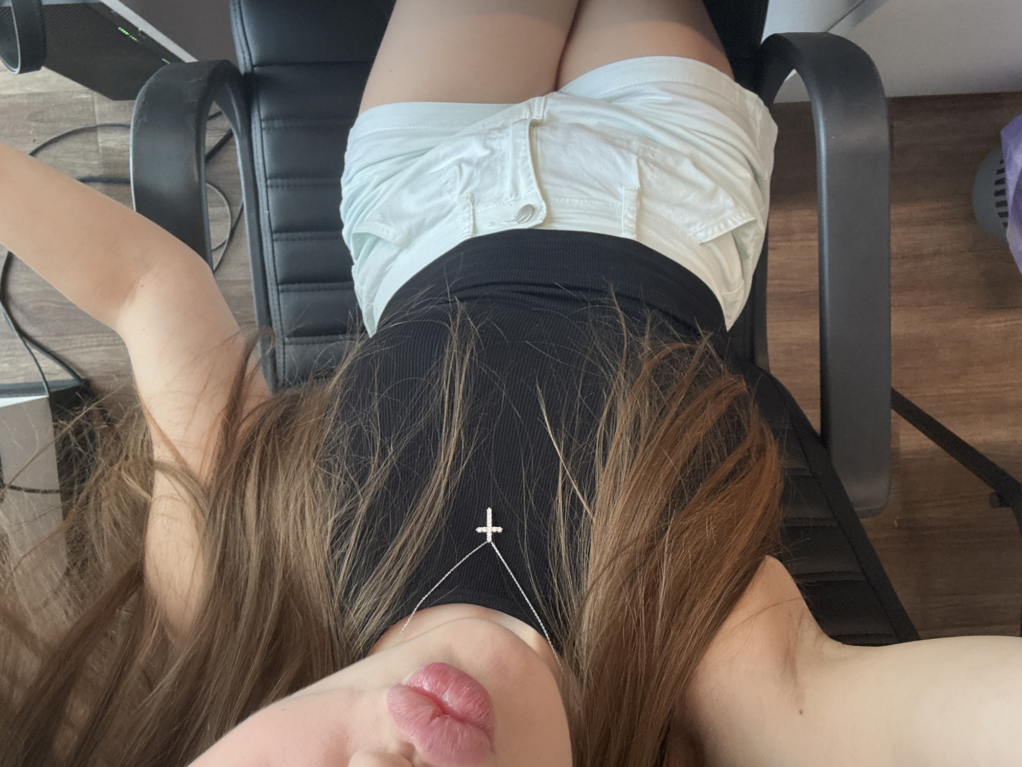A person is lying on a black chair. They are wearing a black tank top and light blue shorts. A silver necklace with a cross pendant is visible around their neck. The person has long, brown hair. The background shows a wooden floor and some electrical cords. There is no information to place this image in Dnipro, Ukraine.