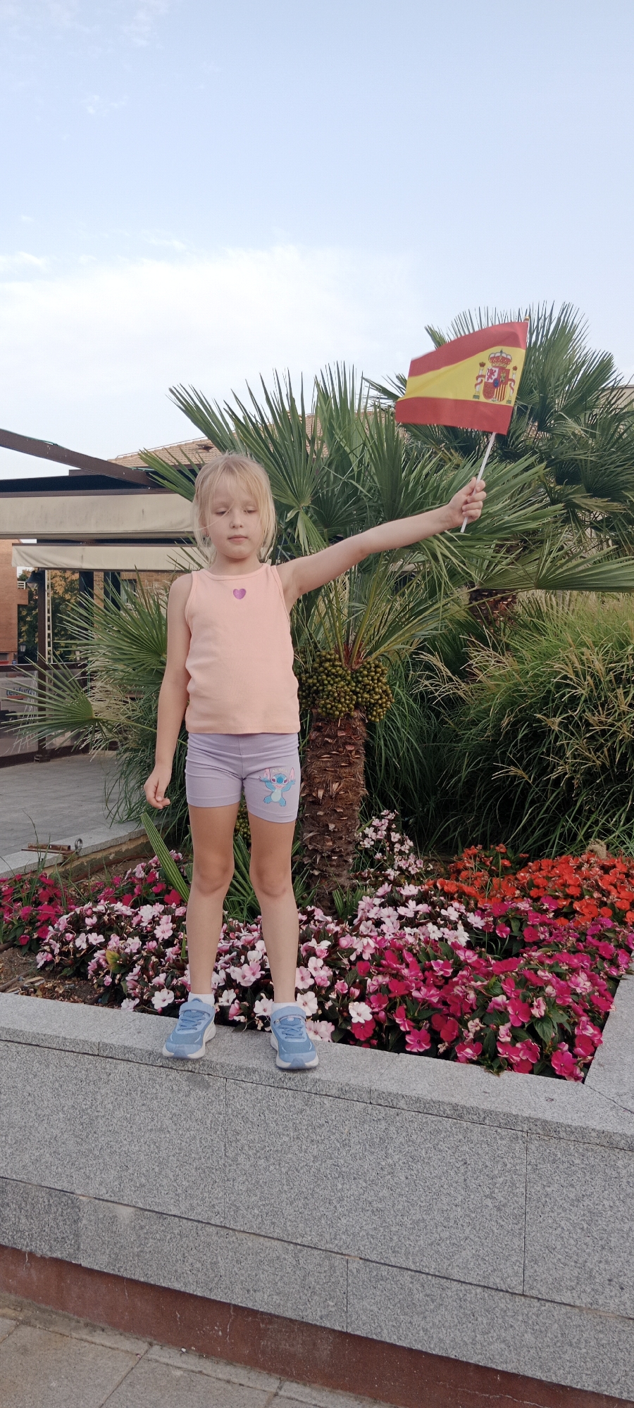 A young Caucasian girl with blonde hair, wearing a peach-colored tank top and light purple shorts with a cartoon character on the left leg, stands on a stone ledge, holding a small Spanish flag on a white stick in her right hand. Her left arm is extended forward, and she is looking down. She is wearing light blue sneakers. Behind her are palm trees, lush green foliage, and a vibrant flower bed filled with pink, white, and red flowers. A building with a tiled roof and awnings is visible in the background. The sky is a pale blue with some light clouds. This scene is located in Galapagar, Spain.