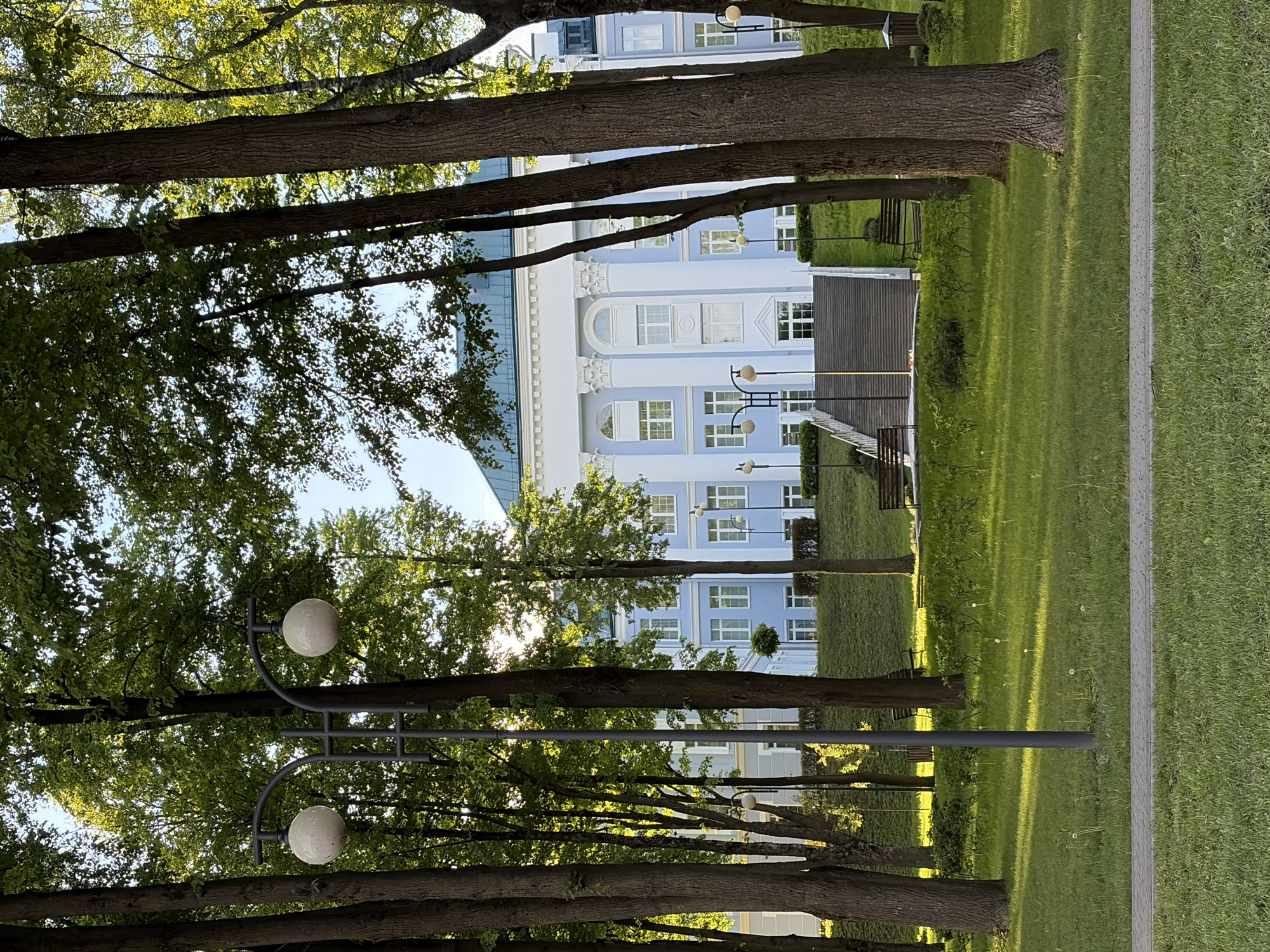 The image shows a view of a building through a line of trees in a park. The building is light blue and white, with multiple windows and architectural details such as columns and arches. Streetlights with globe-shaped lamps are positioned among the trees. The park has a green lawn and a paved walkway along the right edge. The scene appears to be in Minsk, Belarus.