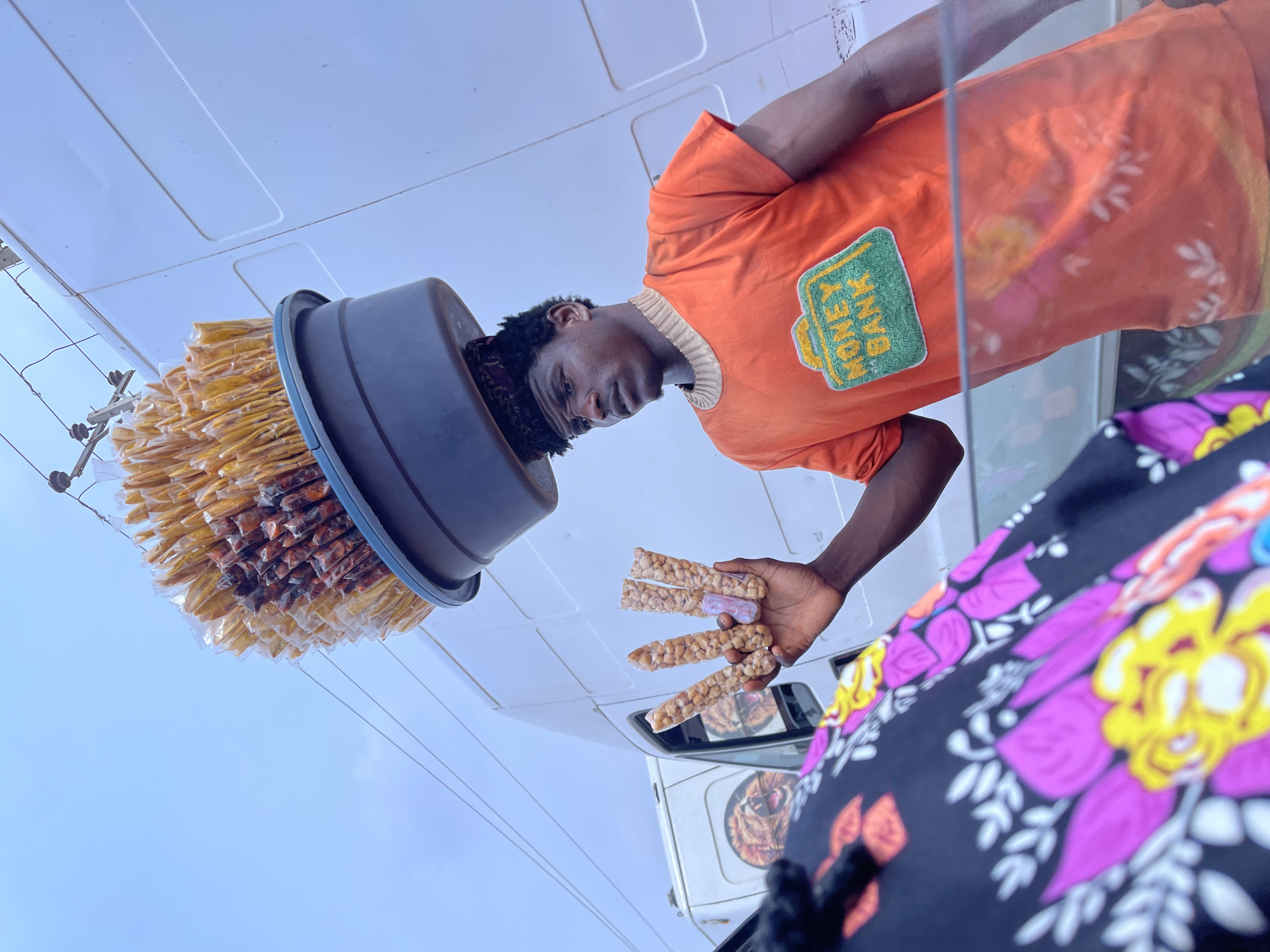 This vertically oriented media file captures a young male street vendor in what appears to be a bright, sunny daytime setting in Ado, Nigeria. The man, with dark curly hair, is positioned centrally, looking downwards. He wears a vibrant orange t-shirt with a light-colored ribbed collar. A prominent green, textured patch on his t-shirt displays the text "MONEY BANK" in yellow lettering. He is also wearing patterned shorts.
On his head, he balances a large, dark-colored basin filled with numerous individually packaged snacks, which appear to be long, thin, and brownish, possibly a type of fried dough or plantain chips. In his right hand, he holds up several lighter-colored, elongated snacks, tightly wrapped in clear plastic, resembling peanut brittle or *kuli-kuli*. These are likely being offered for sale.
The background shows a clear, light blue sky above a white surface, possibly the side or roof of a truck or van. Part of a white vehicle, a truck or van, is visible in the lower background, with reflections showing what looks like a lion's face on its side window. Electrical wires or utility lines are also visible against the sky. A portion of another person, wearing a black garment with bright pink, purple, and yellow floral patterns, is visible in the lower right foreground. The overall scene depicts a typical street vending activity.