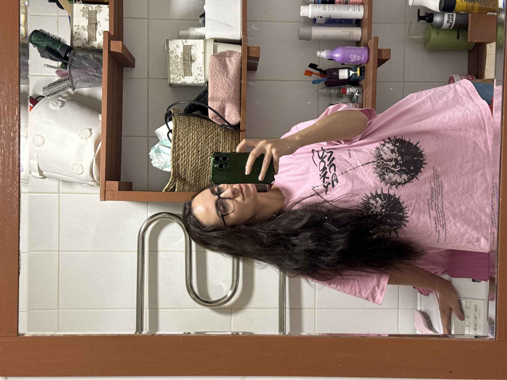 A woman with long dark hair and glasses is reflected in a bathroom mirror. She holds a smartphone in her right hand. The woman wears a pink t-shirt with a black print. The bathroom features white tiled walls, a metal towel rack, and a wooden shelf stocked with toiletries, cosmetics, and storage containers. The location is the city of Krasnoyarsk, Russia.