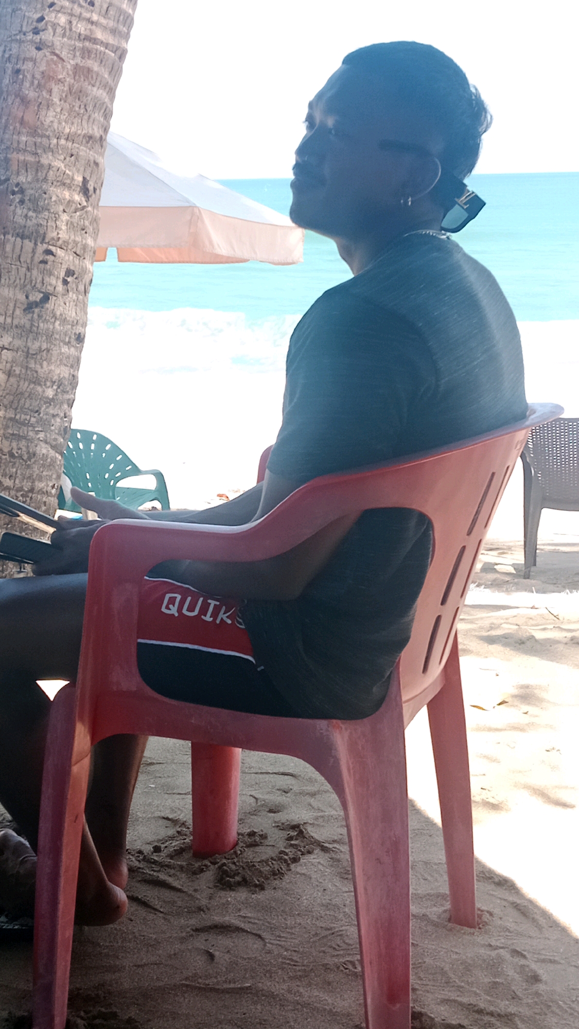 A man sits on a red plastic chair on a beach in Kuta, Indonesia. He is facing to the right, with his back to the camera. He is wearing a dark t-shirt and dark shorts with a red and white logo that says "QUIKSILVER". He has sunglasses perched on the back of his head. The man is sitting next to a palm tree. In the background, there is a beach umbrella and the ocean. The sand is light brown and there are footprints in the sand.
