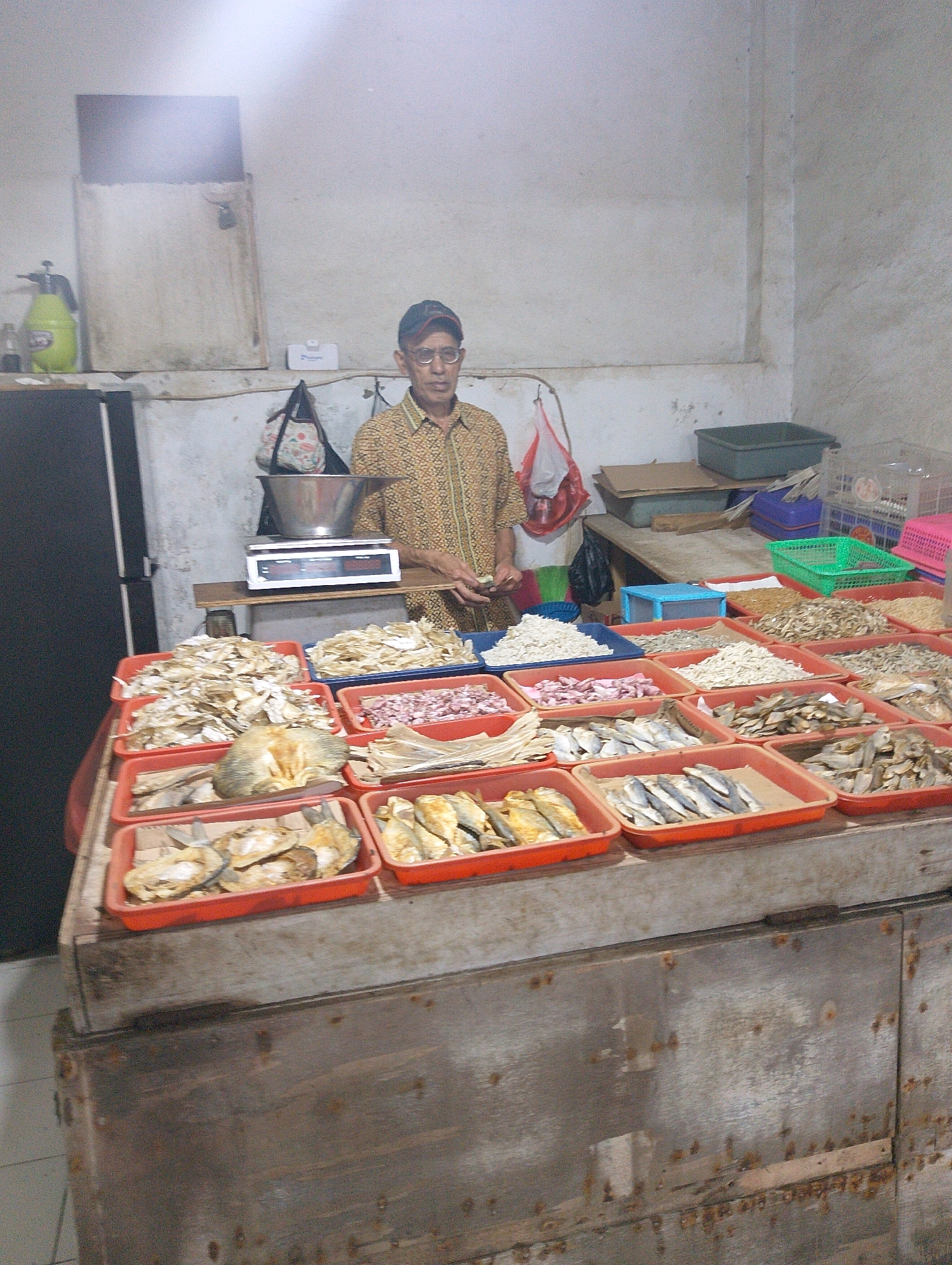 The image depicts a man wearing glasses and a patterned shirt standing behind a stall piled high with various types of dried fish. The stall is located in Lembang, Indonesia, as indicated by the context. The man appears to be a vendor, as he is positioned behind the merchandise. The stall is organized with trays of dried fish, sorted by type, and a scale is visible on a wooden surface behind the man, suggesting it is used for weighing purchases. The environment appears to be an indoor market or shop, with a rough-textured wall in the background and a tiled floor. Other market items, such as baskets and containers, are also visible. The lighting is somewhat dim, contributing to the market's atmosphere.