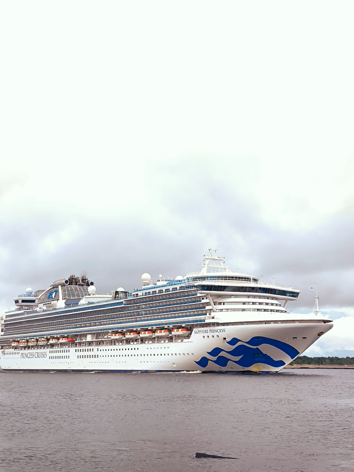 A large white cruise ship, named "SAPPHIRE PRINCESS" and also displaying "PRINCESS CRUISES" on its hull, is sailing on dark, choppy water. The ship has multiple decks with rows of windows, and lifeboats are visible along its side. The bow of the ship features a distinctive blue wave-like design. The sky is overcast with grey clouds. In the background, a treeline is faintly visible, suggesting land. The scene is likely a port or a waterway near a city, possibly during daytime given the visible details. Without further information or context, pinpointing the exact location in Riga, Latvia, is not possible based solely on the visual elements present.