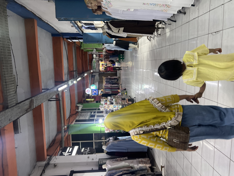 An indoor marketplace displays a woman with a yellow hijab, patterned jacket, and jeans, holding hands with a child in a matching yellow dress. Merchandise including clothing and accessories lines the pathway. Storefronts are visible along the sides, with mannequins displaying garments. Overhead, cables and lighting fixtures illuminate the space. The marketplace is bustling with activity and offers a variety of shopping options.
