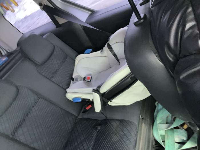 The scene depicts the interior of a vehicle, specifically the rear passenger compartment, located in the city of Podvyaznovo, Russia. A light-colored, unoccupied infant car seat is secured on the rear passenger seat directly behind the front passenger seat. The rear seats feature dark grey fabric upholstery with a quilted or textured pattern. The back of the front passenger seat, upholstered in black material, is partially visible on the right. In the immediate foreground, a segment of a person's upper body or arm, wearing a black puffy jacket, is visible. On the vehicle floor in the foreground, a bundle of light blue or teal webbing straps is present. Through the rear window, a blurry, bright, light-toned exterior environment is visible, consistent with an overcast sky or snowy conditions. No individuals are depicted occupying the car seat or directly interacting within the visible frame.