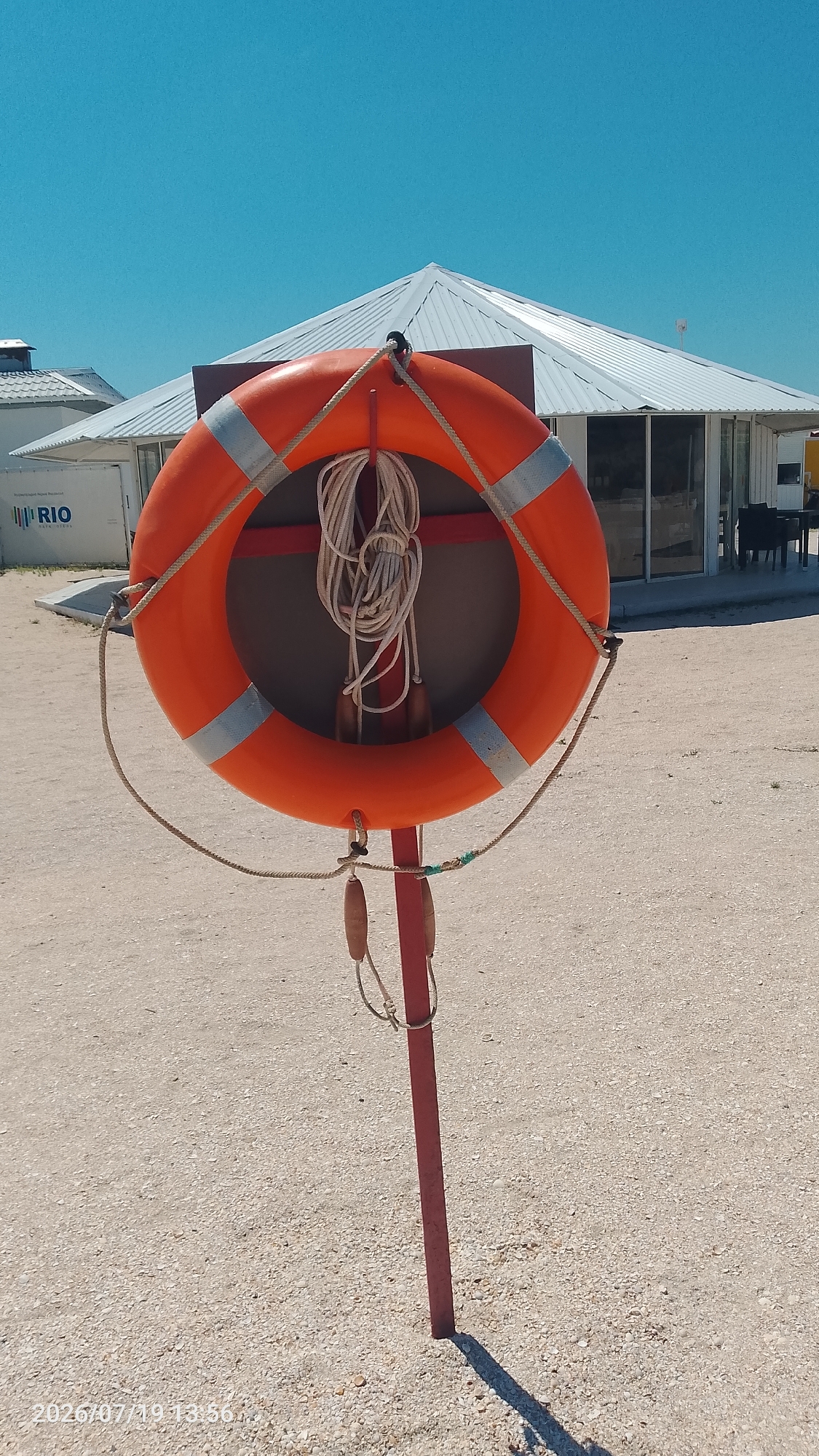 A bright orange lifebuoy with reflective silver bands is mounted on a red metal pole on a sandy beach. The lifebuoy has a coiled rope attached to it, with two wooden handholds hanging from the rope. In the background, there is a white building with a multi-peaked roof, which appears to be a beachside establishment. The sky is clear and blue. The lower left corner of the image has a timestamp "2026/07/19 13:56". The location is identified as Мысовское сельское поселение, Ukraine.