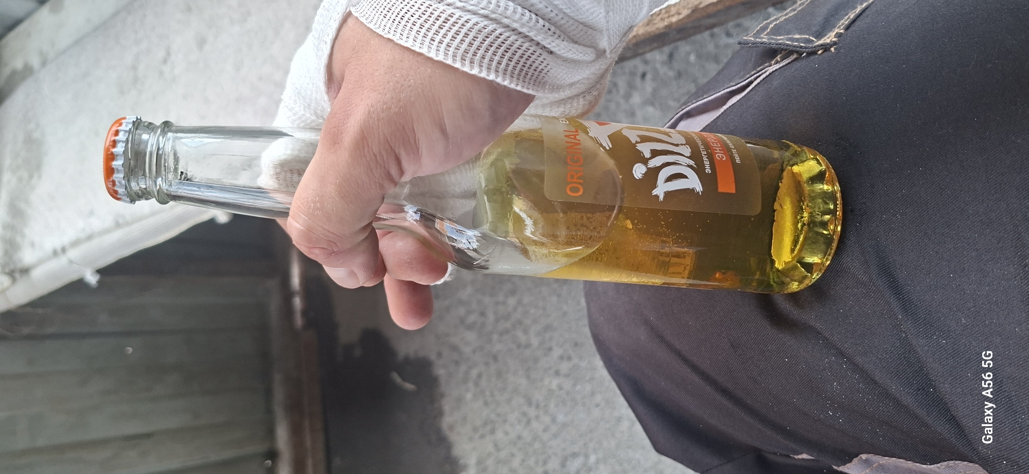 A hand wearing a white bandage is holding a glass bottle of Dizzz Original energy drink. The bottle contains a yellow-green liquid and has a metallic orange cap. The label on the bottle is brown with orange and white text. The person is wearing dark clothing, possibly pants. The background is out of focus and appears to be an outdoor setting with a corrugated metal wall and a white structure overhead. The bottom left corner of the image displays "Galaxy A56 5G". The scene does not provide any specific indicators of the city of Almaty, Kazakhstan.