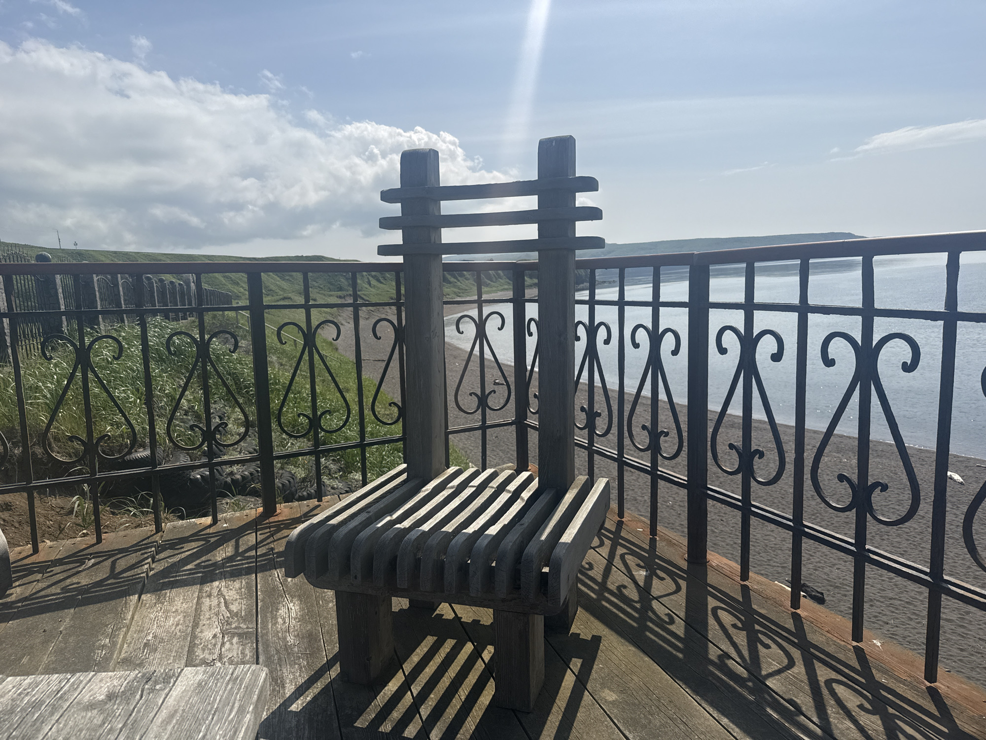 A wooden bench with a high, slatted back is positioned on a wooden deck overlooking a beach and the ocean. The bench is made of weathered wood and appears to be handmade. The deck is also made of wood planks and has shadows cast on it from the bench and a decorative metal railing. The railing, which features heart-shaped scrollwork, separates the deck from a grassy embankment. Beyond the embankment, a dark-colored beach stretches along the coastline. The ocean is calm and blue, with a faint horizon line visible. The sky is bright blue with scattered white clouds. The overall scene suggests a scenic viewpoint, possibly a park or a coastal lookout. This location is in Kuril'sk, Russia.