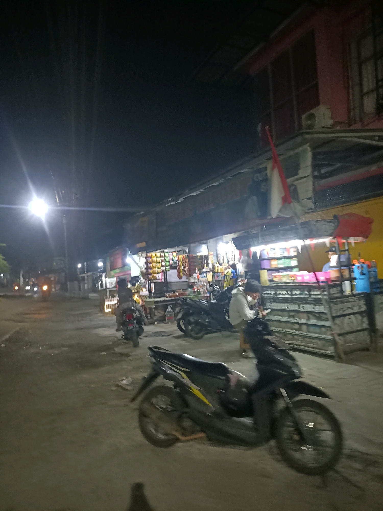 This is a nighttime photograph taken on a street in Wanajaya, Indonesia. The scene depicts a small street market with several stalls set up. In the foreground, a black motorcycle is parked on the dirt road. Further down the street, other motorcycles are visible, some with riders. One stall displays an array of colorful packaged goods. A red and white flag is mounted on a pole above one of the stalls. The buildings lining the street appear to be two stories high, with some windows and an air conditioning unit visible. The overall lighting is dim, with the exception of bright streetlights and the lights from the stalls, creating a somewhat hazy atmosphere.