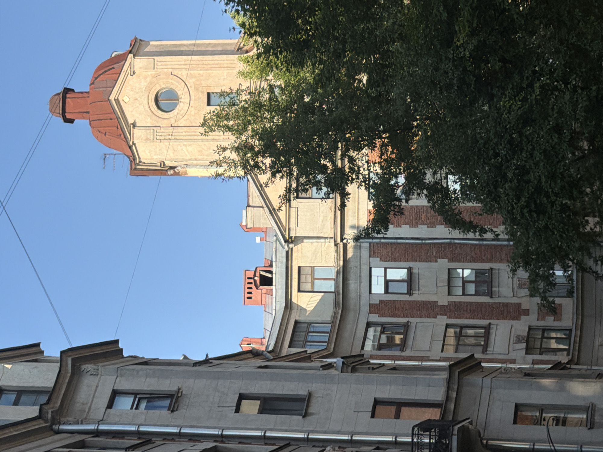 This is a medium-shot, low-angle, vertically oriented photograph of buildings and a tree against a clear blue sky. The sun appears to be shining, suggesting daytime, likely morning or afternoon given the brightness of the sky and the light on the buildings.
The main subjects are parts of several buildings, seen from a low vantage point looking upwards. The building on the left has a distinctive curved, reddish-brown roof with a small turret or spire. Below this roof is a light-colored section of the building with a round window. Further down, the facade has a more modern, grey material, with multiple rectangular windows. Another building to the right features a more traditional brick facade with cream-colored decorative elements and many rectangular windows. The topmost section of this building is obscured by a large, leafy green tree. The lower parts of the image show the grey, sloped roofs of more buildings with gutters and more rectangular windows. The overall architectural style appears to be a mix of older, possibly Art Nouveau or similar, European styles and more utilitarian modern additions.
There are no people visible. The objects present are architectural elements like windows, roofs, chimneys, and a metallic gutter. A prominent tree with dark green foliage occupies the upper right portion of the frame, partially obscuring the buildings behind it. Several thin black wires are stretched across the sky, indicating the presence of utility lines.
No activity or event is taking place; it is a static scene of urban architecture. No text is visible in the image. The scene evokes a sense of urban life and the layered history of city development.