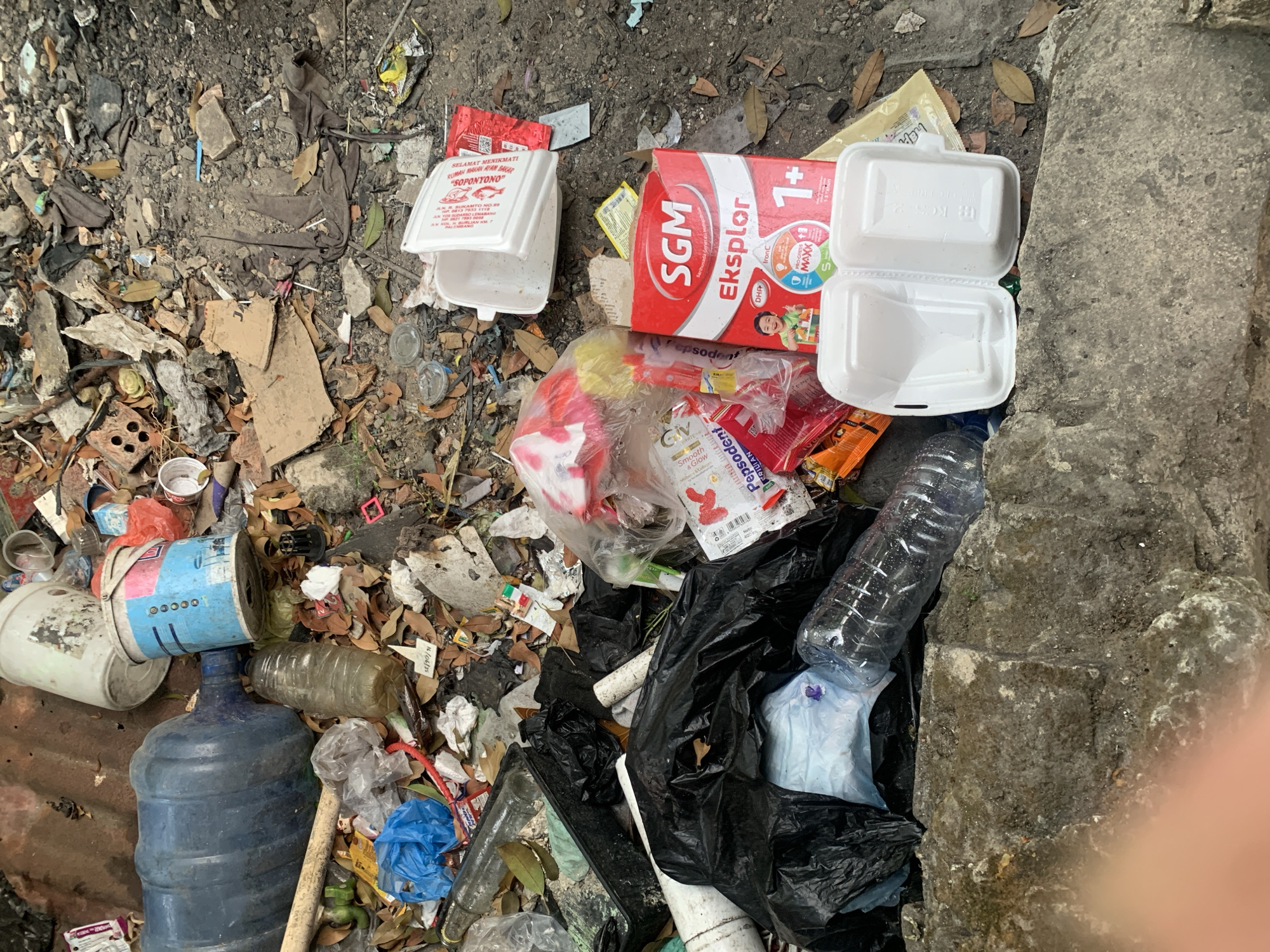 In Palembang, Indonesia, a pile of uncollected garbage lies scattered on a dusty, uneven ground. The debris includes various plastic items such as a large blue water jug, several plastic bottles, a blue plastic bag, and white styrofoam containers. Cardboard pieces and broken bricks are also visible amongst the waste. A prominent red and white box of "SGM Eksplor" formula milk is present, along with smaller packaging items for products like Pepsodent toothpaste. The scene suggests a general area of neglect and accumulation of waste.