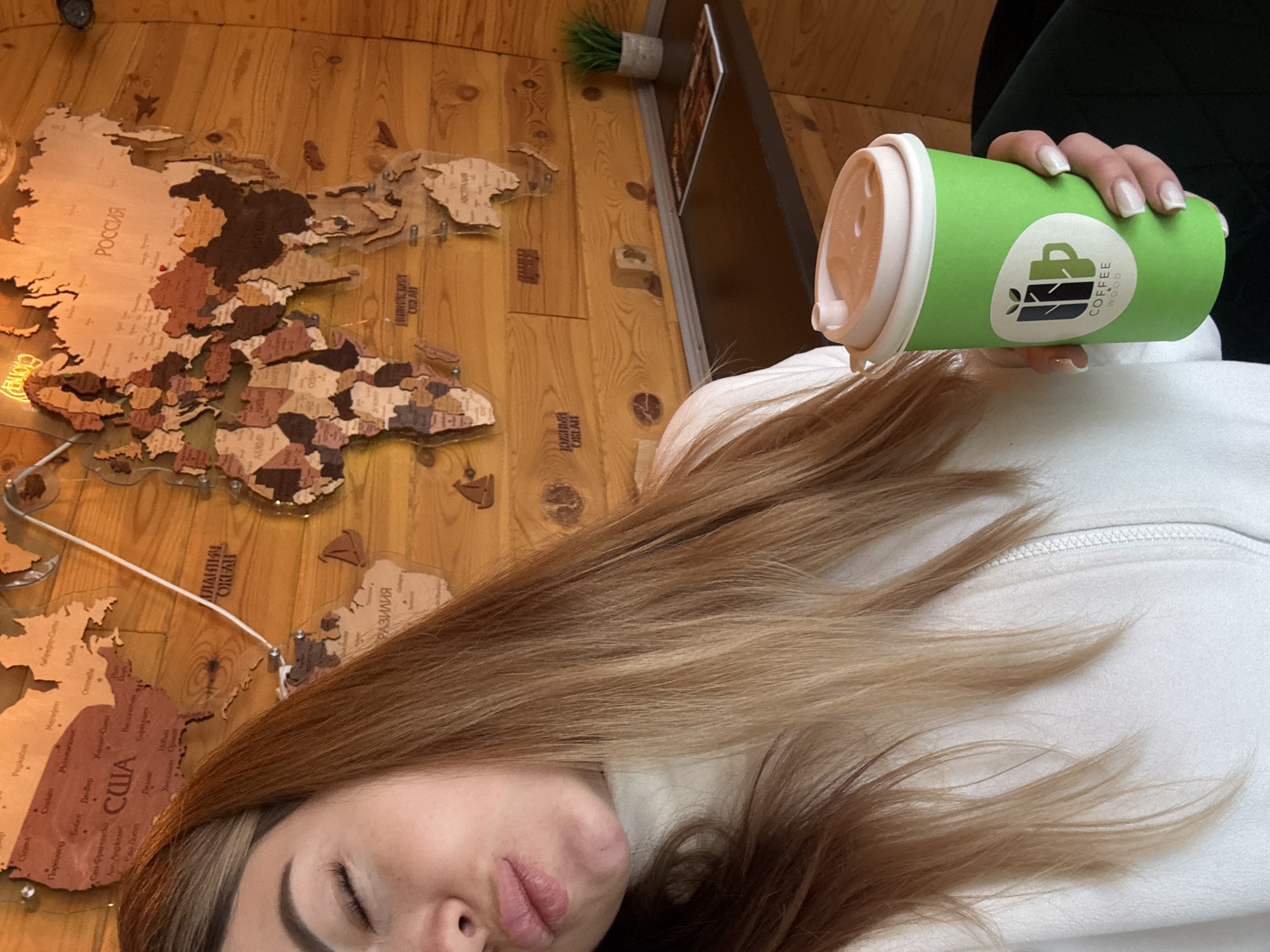 A low-angle, slightly canted, indoor shot depicts a person lying down, holding a cup of coffee in their right hand. The person has long, light brown hair that is spread out on a white zippered garment. Their eyes are closed, and their lips are slightly puckered.
The coffee cup is green with a white lid and features a logo that reads "COFFEE WOOD" alongside an image of coffee beans. The person's hand with manicured nails holds the cup.
The background consists of a wooden wall featuring a decorative world map. The map shows outlined countries, including "РОССИЯ" (Russia) and "США" (USA). A white cable runs across part of the map. To the right, a green artificial plant is mounted on the wall above a dark shelf. The location is Unknown, Unknown.