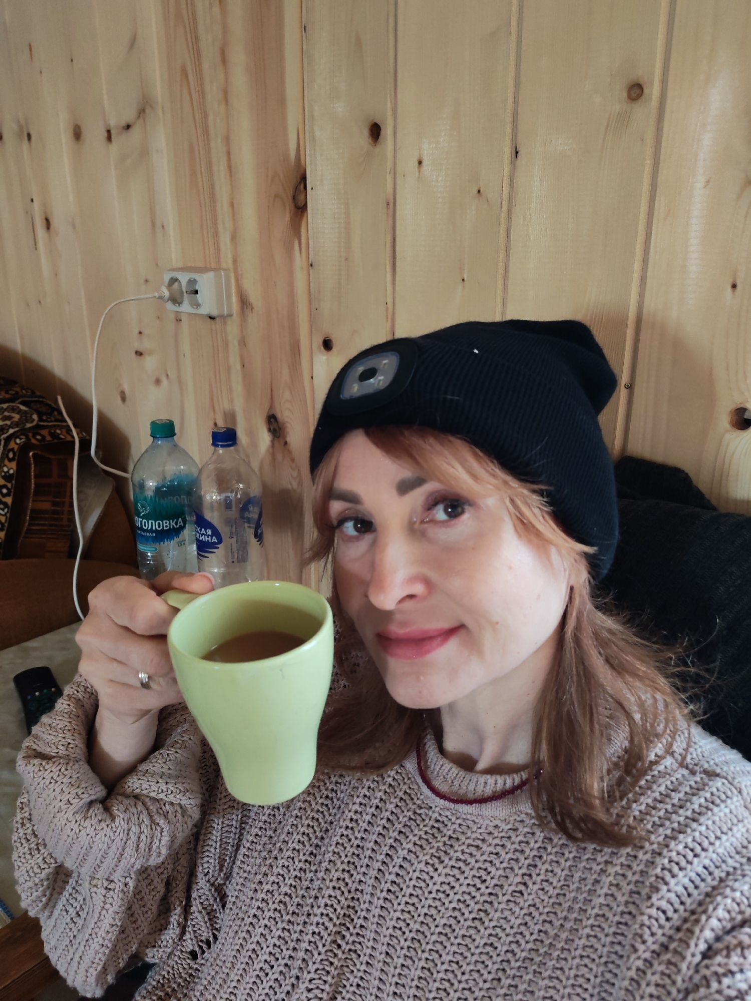 A woman is holding a cup of coffee, wearing a light pink sweater and black knit hat with a light on the front. Behind her are two water bottles on a shelf, and a power outlet is mounted on the wooden wall. The bottles have Russian labels on them: "Оголовка" and "СКАЯ ЖИНА". The background appears to be a rustic wooden interior. It is not possible to verify the location without additional information.