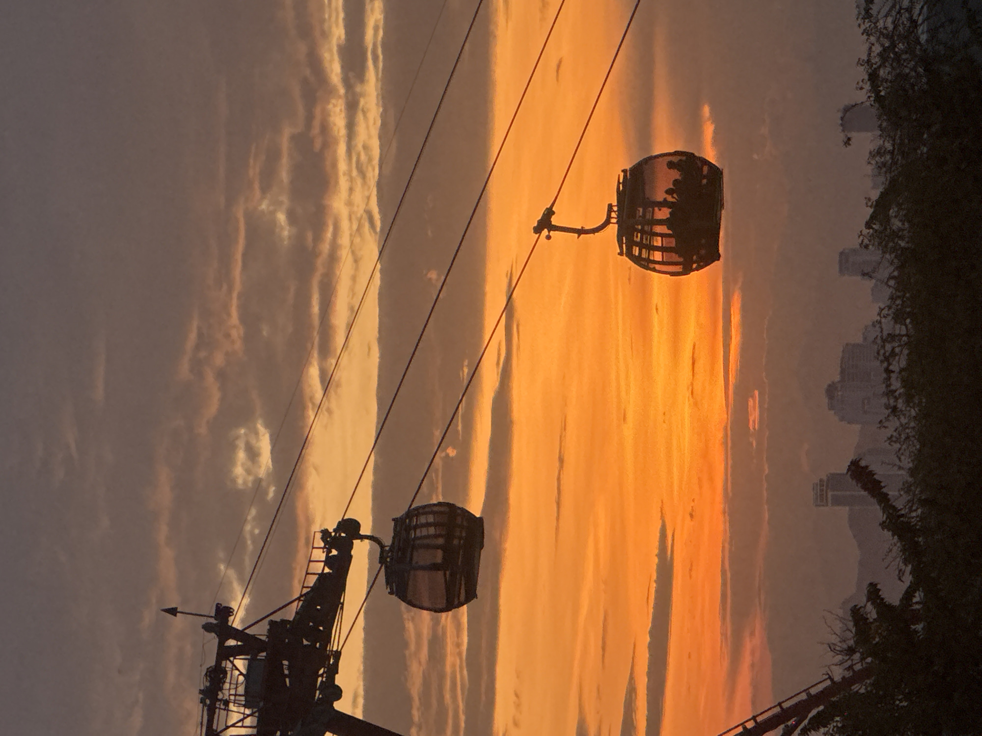 The image shows two cable cars silhouetted against a vibrant orange and purple sunset sky. The cable cars are suspended from cables, with one car closer to the foreground on the right and another car further in the distance on the left. Both cars appear to be filled with passengers. In the background, a hazy cityscape with buildings and mountains is visible. The foreground is occupied by dark silhouettes of trees and foliage. This scene is characteristic of the cable car system that connects Hon Tre Island to the mainland in Nha Trang, Vietnam, offering panoramic views of the city and the bay, especially during sunset.