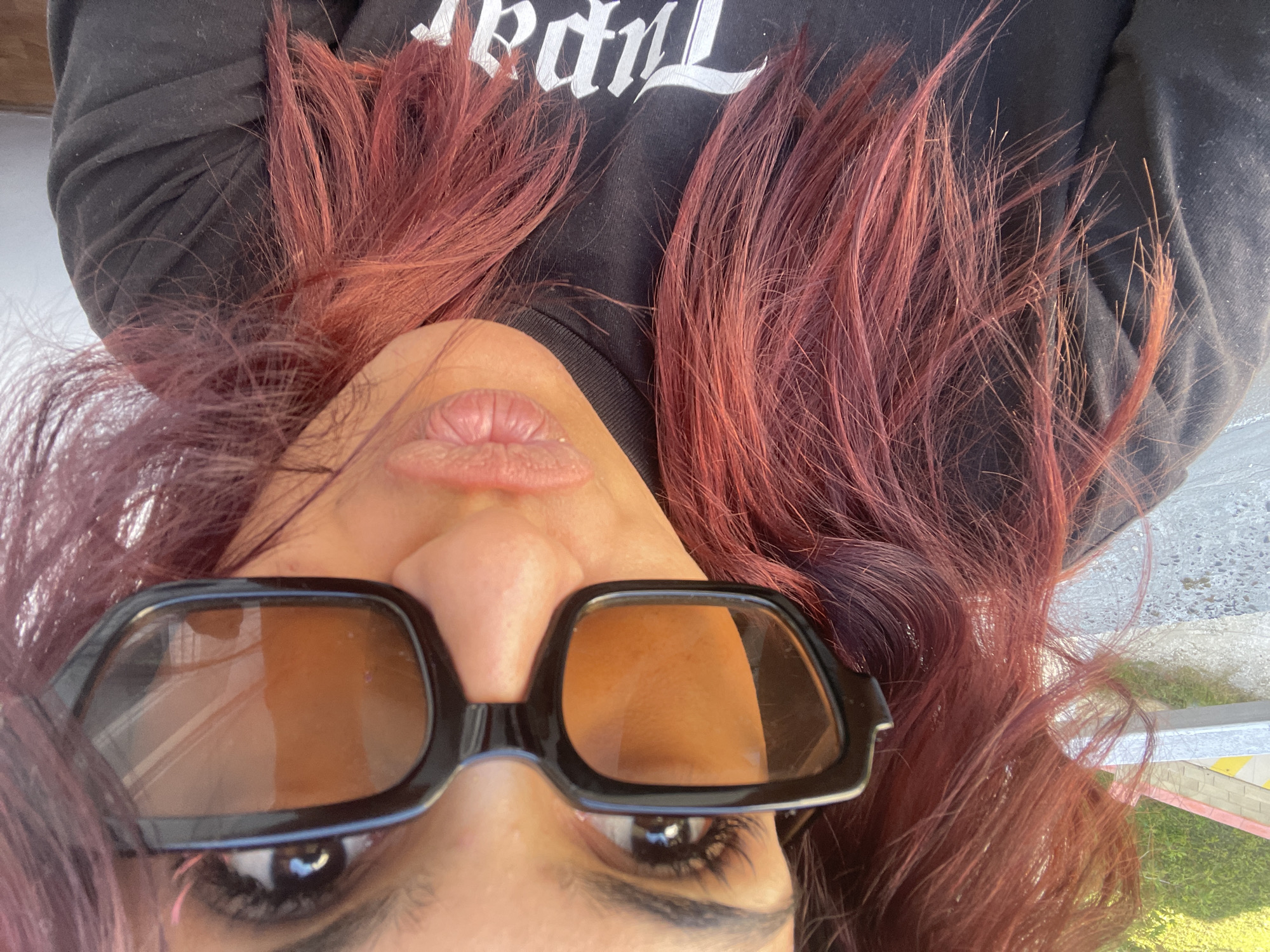 The image shows a person from an upside-down perspective, wearing dark sunglasses with brown-tinted lenses and a black shirt with white gothic-style lettering. The person has light skin, full lips, and dark eyelashes. Their hair is dyed a reddish-brown color and is spread around their face. The background is blurred, showing what appears to be a street or outdoor area, possibly in a city, with indistinct colors suggesting buildings and greenery.