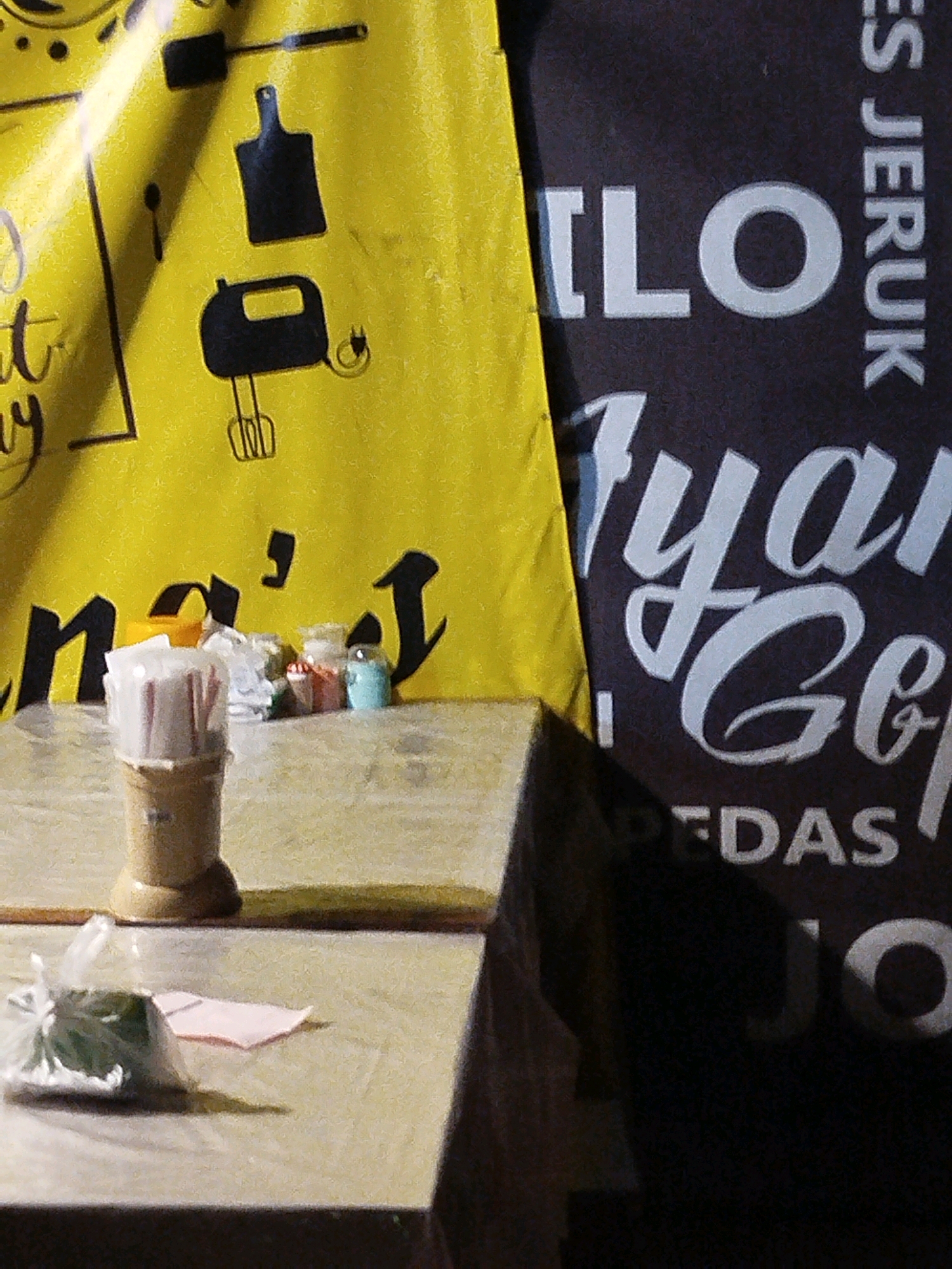 The image shows a close-up view of a table with various items on it, set against a backdrop of two large banners. The banner on the left is bright yellow with black graphics of kitchen utensils, including a spatula, a chopping board, and a hand mixer. The banner on the right is dark with white lettering in a stylized font. Some of the visible text includes "ES JERUK", "ILO", "ryan G&", and "PEDAS".
On the table, in the foreground, is a small beige cup holding several pink items that appear to be paper or straws. To the left of the cup, a clear plastic bag contains green leafy material, possibly herbs or vegetables, and a folded pink piece of paper is visible underneath. In the background, on the table, are several small containers. One appears to be an orange lid, and next to it are two small jars. One jar contains a red substance, possibly spices or candy, and the other contains a light blue granular substance. The table surface is textured and appears to be made of wood or laminate. The lighting suggests an outdoor or semi-outdoor setting, possibly a street food stall or a small shop, given the banners and the arrangement of items. The specific location in Mojopanggung, Indonesia, is not visually evident from the image itself, but the text on the banners might refer to local products or businesses.