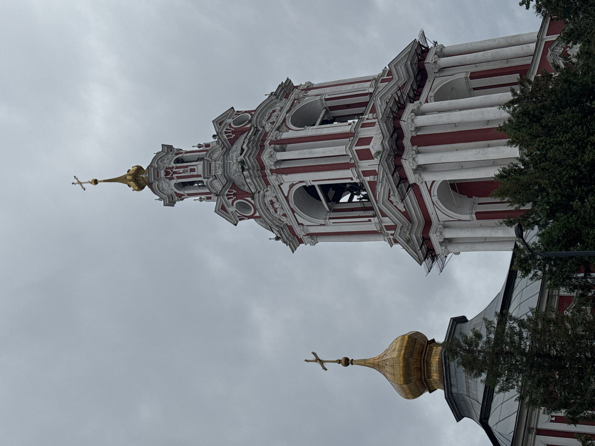 The image features a tall, ornate bell tower, likely part of a religious structure in Moscow, Russia. The tower has a distinctive red and white striped pattern and is built in a baroque style with multiple tiers and decorative elements. At the top is a golden cross.
The lower portion of the image includes a golden dome with a cross, suggesting another part of the same or an adjacent church. Parts of the building and a tree block a full view of the dome.
The sky is overcast, creating a dull light. The image is shot from a low angle, emphasizing the height of the bell tower.