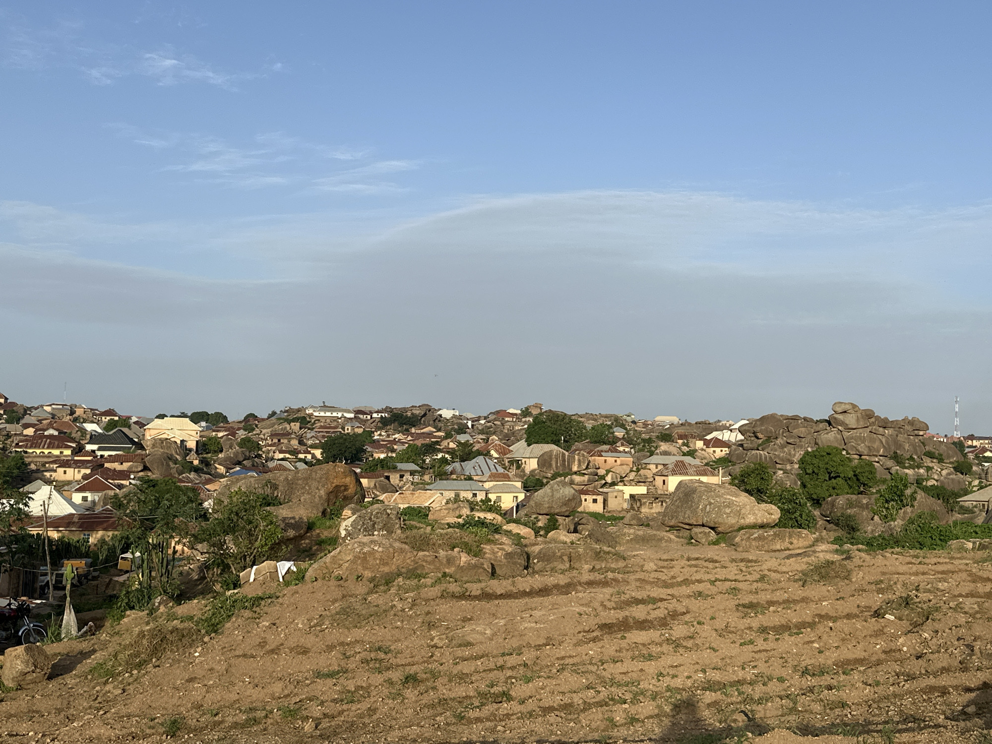The image depicts a sprawling settlement in the city of Jos, Nigeria, characterized by its unique integration with rocky outcrops. The foreground consists of a barren, reddish-brown earth terrain with subtle furrows suggesting agricultural activity. A large, weathered boulder dominates the lower left of this area, with smaller rocks and sparse dry vegetation scattered throughout.
Moving towards the midground, the landscape transforms into a densely populated residential area built amongst and around significant rock formations. Numerous houses with corrugated metal roofs, varying in color from red and brown to grey and blue, are visible. The architecture appears to be a mix of traditional and modern styles. Green trees and shrubs sprout from crevices in the rocks and between buildings, adding a touch of nature to the urban environment. Some white laundry is draped over a large rock in the mid-right portion of the frame. A few vehicles, including what appears to be a motorcycle on the far left, are present.
The background reveals a continuation of the settlement extending over undulating terrain, further punctuated by rocky hills. The sky is a clear, pale blue with wispy white clouds, suggesting it is either early morning or late afternoon. The overall impression is a vibrant community living in harmony with its striking geological surroundings.