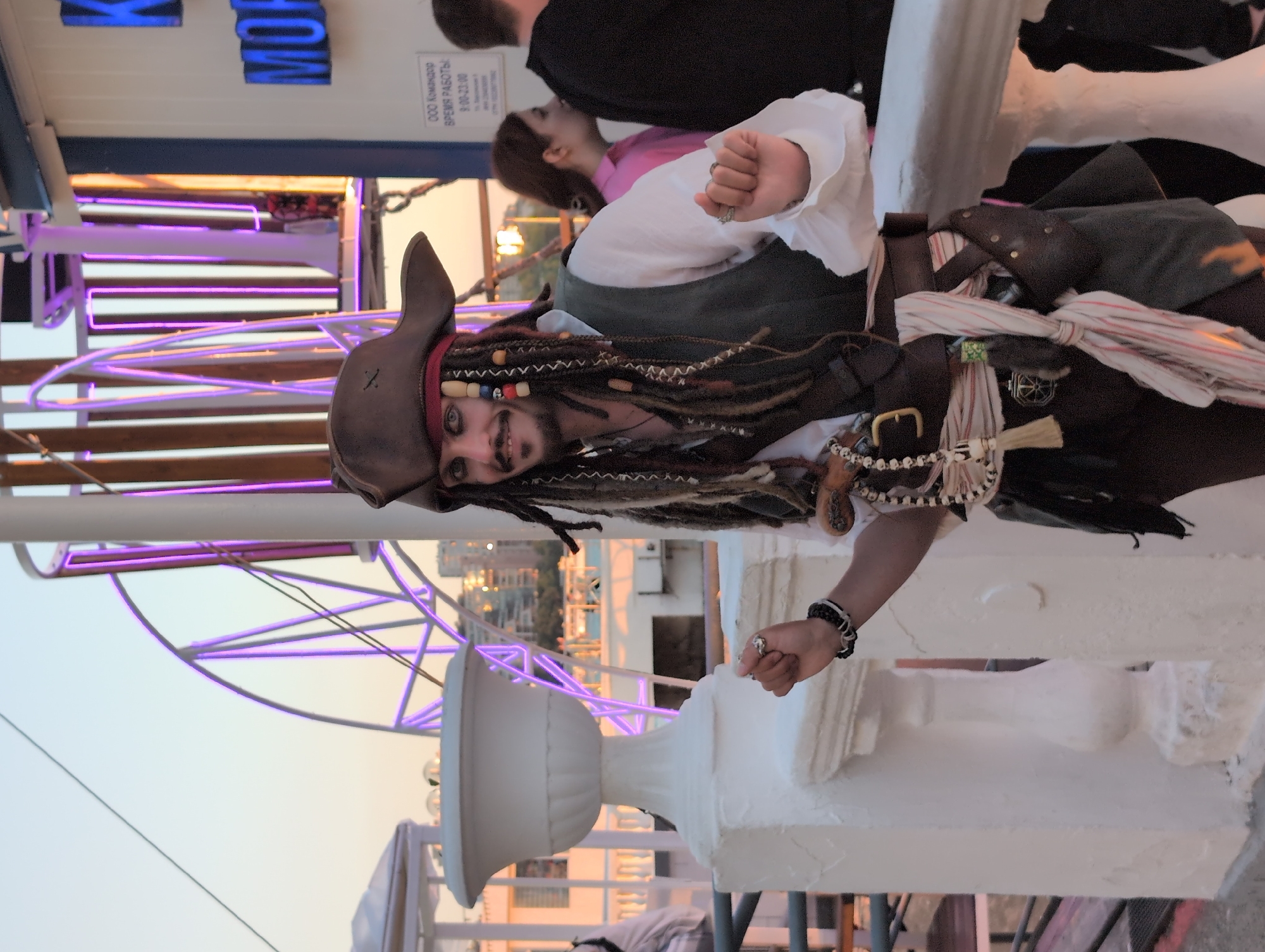 A man dressed as Captain Jack Sparrow poses for a photograph on a boat in Gelendzhik, Russia. The man wears a tricorn hat, dreadlocks, eye makeup, a white ruffled shirt, a vest, and a sash. He holds his left fist up and his right hand is partially visible near his waist. In the background, a Ferris wheel with purple lights is visible, along with a building with blue lettering that reads "MOP". The scene appears to be on an outdoor deck, possibly a pier or a boat, with a white railing and urns. The time of day is late afternoon or early evening, as indicated by the soft, golden light.