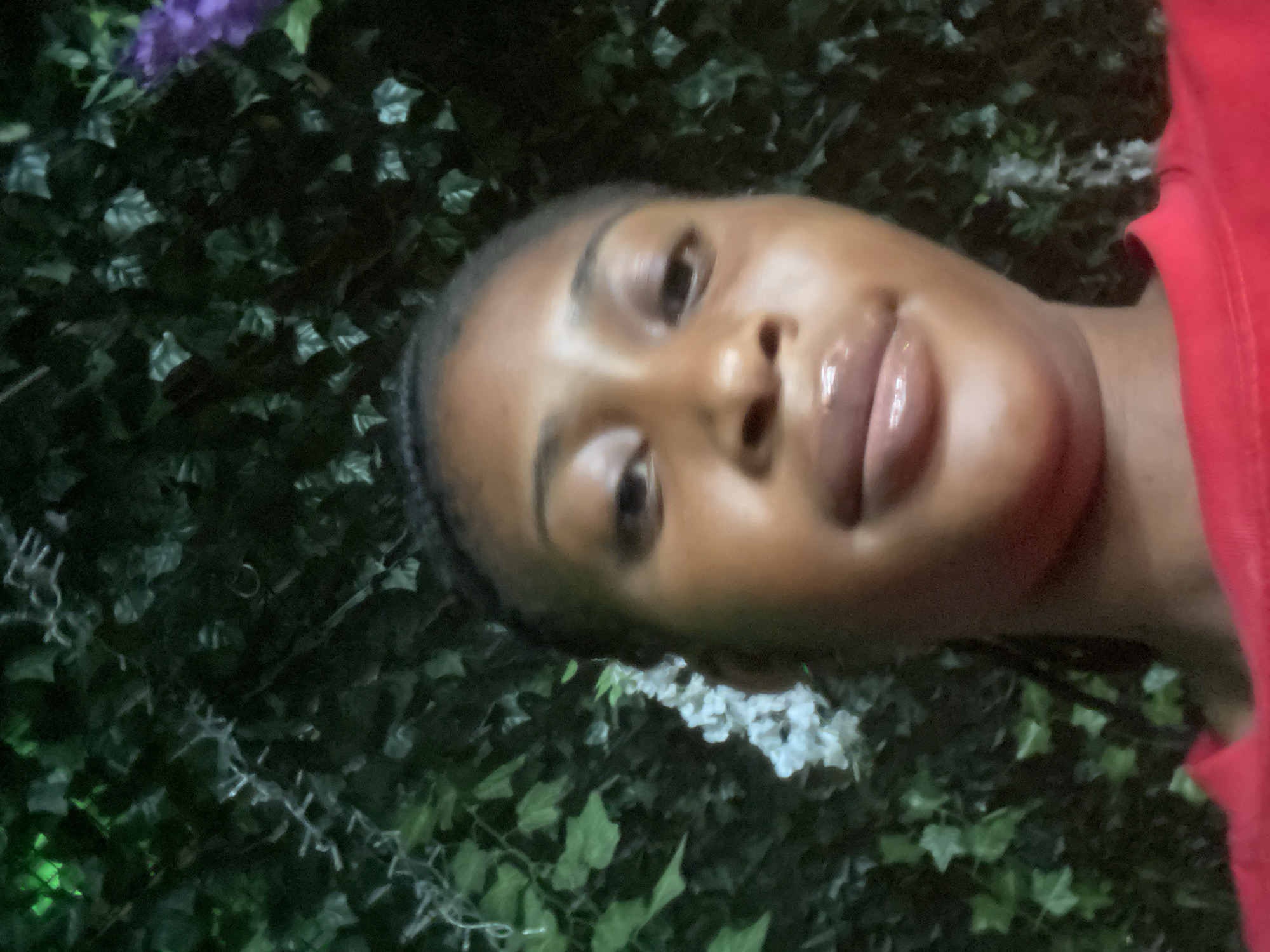 The image is a close-up, slightly blurry, selfie of a young Black woman with braided hair. She is wearing a red shirt. Her lips are glossy, and she appears to be looking directly at the camera. Behind her, there is a dense, dark green backdrop of artificial foliage, possibly ivy, with some white flowers and hints of purple on the right side. The lighting is dim, suggesting an indoor or nighttime setting, possibly in a garden or event space. There is no specific identifiable location or context to determine it is in Abuja, Nigeria.