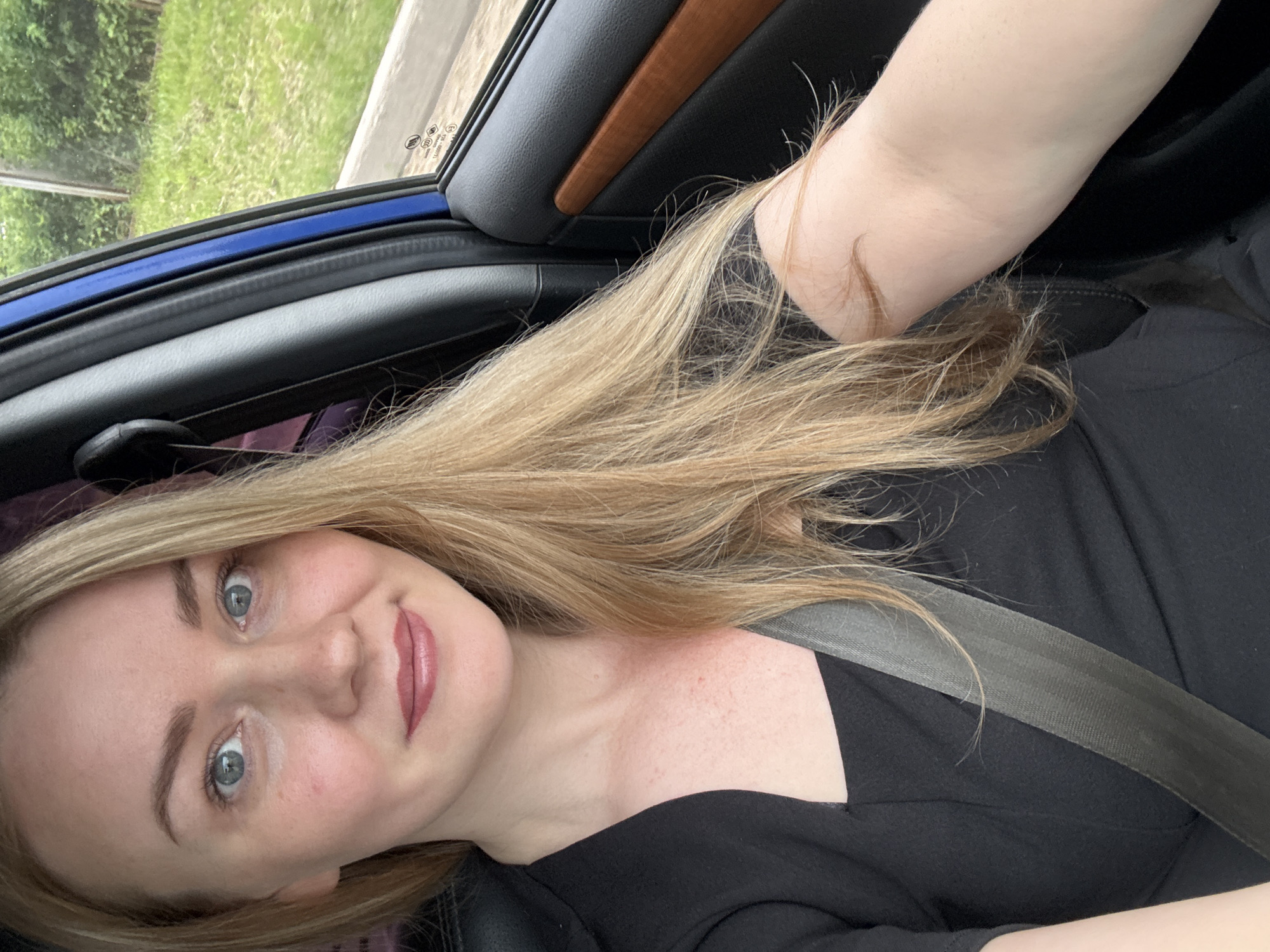 A blonde woman is in the passenger seat of a car, facing the camera. She is wearing a black dress and a seatbelt. The car's interior is visible, including the blue door trim and a wooden accent. Outside the window, lush green trees and grass are visible, suggesting a rural or suburban setting. The image appears to have been taken during the daytime, with natural light illuminating the scene. There is no indication of the specific location in Chaikovsky, Russia, other than the implied context of the prompt.