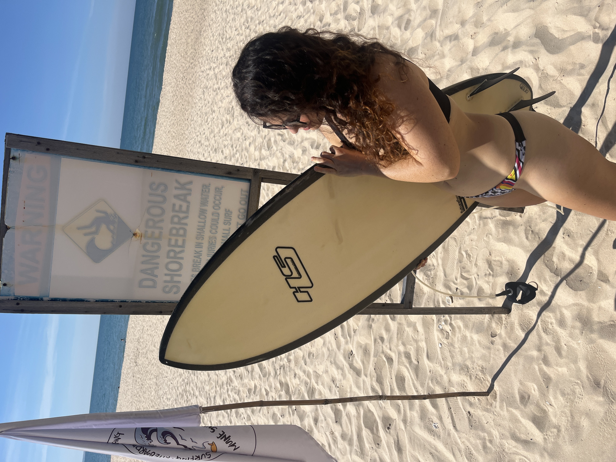 A woman with long, curly brown hair is standing on a sandy beach holding a surfboard. She is wearing sunglasses and a black bikini. A warning sign that reads "DANGEROUS SHOREBREAK" is behind her. The ocean is visible in the background. The scene takes place on a sunny day.