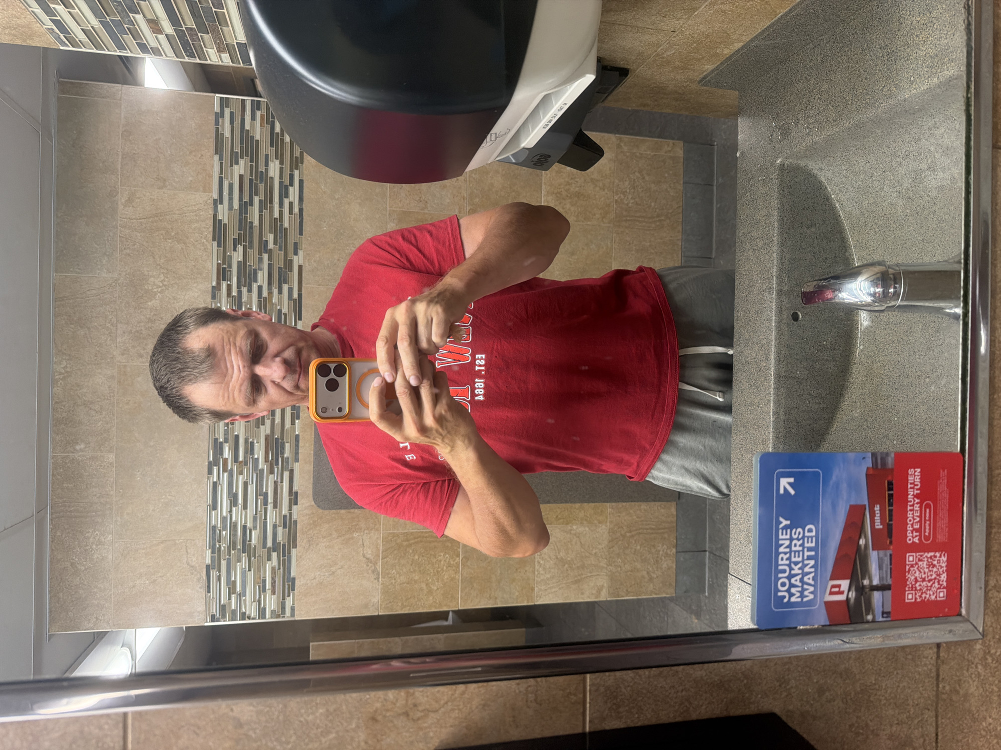 A man is taking a mirror selfie in a public restroom. He is wearing a red t-shirt and grey sweatpants, holding a smartphone in his right hand. In the reflection, the restroom features beige tiled walls, a patterned tiled accent wall, a sink with a chrome faucet, and a black and white paper towel dispenser. On the left side of the mirror, a "JOURNEY MAKERS WANTED" poster with a QR code and a Pilot gas station image is visible. The man's face is slightly obscured by his phone. The setting appears to be a truck stop or rest area, possibly in Lebanon, United States of America, given the context of the "Pilot" branding.