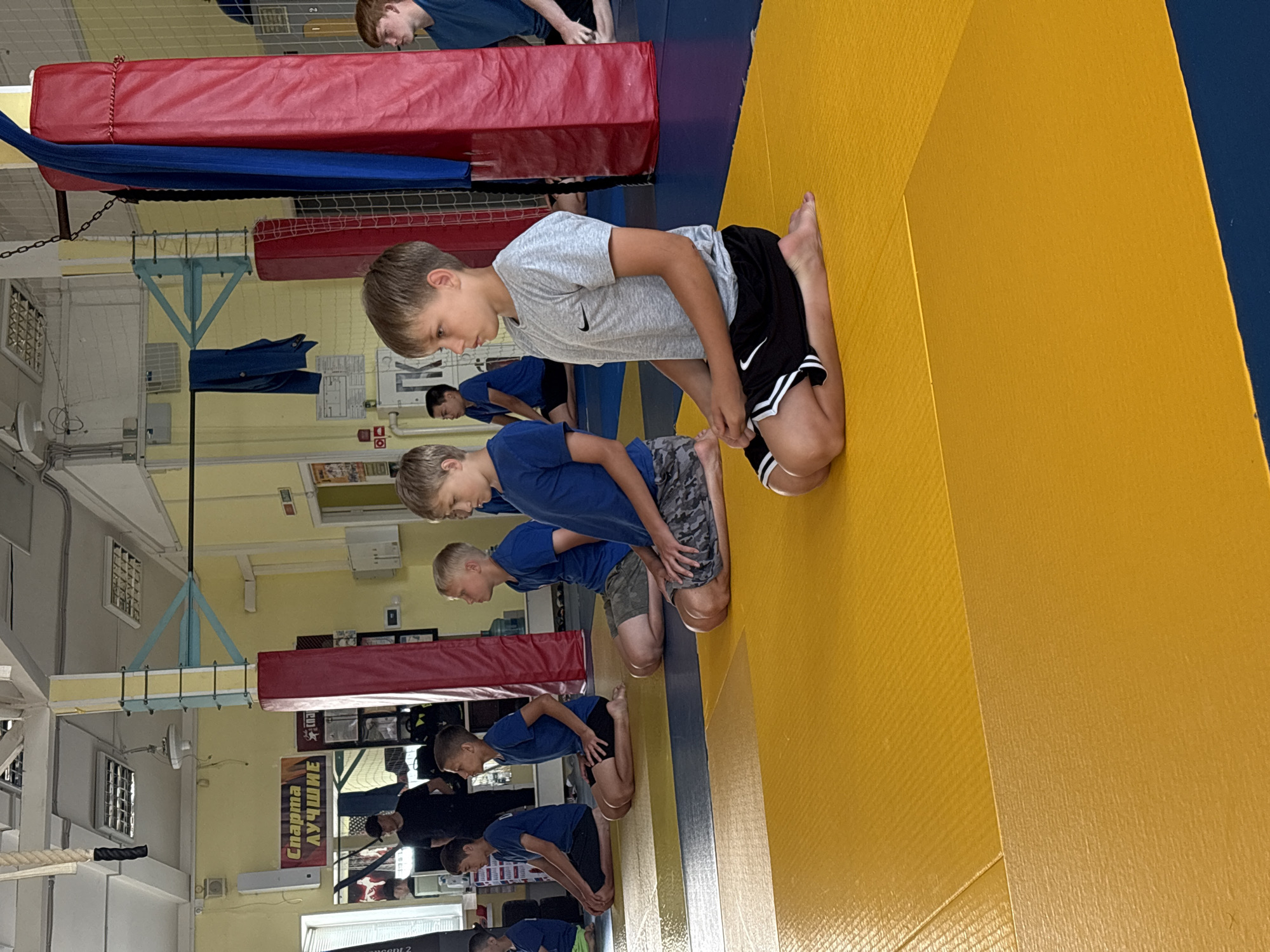The image shows a group of young boys sitting on a yellow mat in a gymnasium. They are all wearing athletic clothing, and most of them appear to be of similar age, around 8-12 years old. Some of the boys are wearing blue t-shirts and shorts, while others are in grey t-shirts and black shorts. They are all sitting in a cross-legged position, with their hands resting on their laps.
In the background, there are several objects visible, including a red padded column, a blue fabric hanging from a metal structure, and some posters on the wall. A man in a black t-shirt and baseball cap is standing in the far left of the image, facing away from the camera. The gymnasium appears to be a martial arts or judo training facility, given the mats and the overall setup.
The image was taken in the city of Chaikovsky, Russia. The overall lighting suggests it is daytime, and the environment appears to be indoors. The boys are likely participating in a training session or a class.