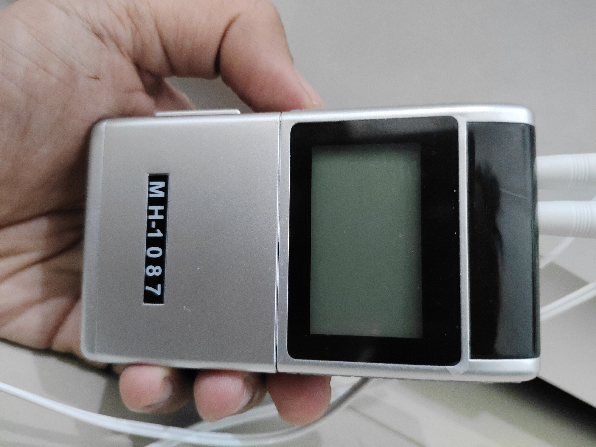 A close-up shot shows a person's hand holding a silver electronic device with a digital display and a number counter. The number counter reads "MH-1087". The device has two white cords plugged into its right side. The background is blurred and appears to be a light-colored surface. The image was taken in Semambung, Indonesia.