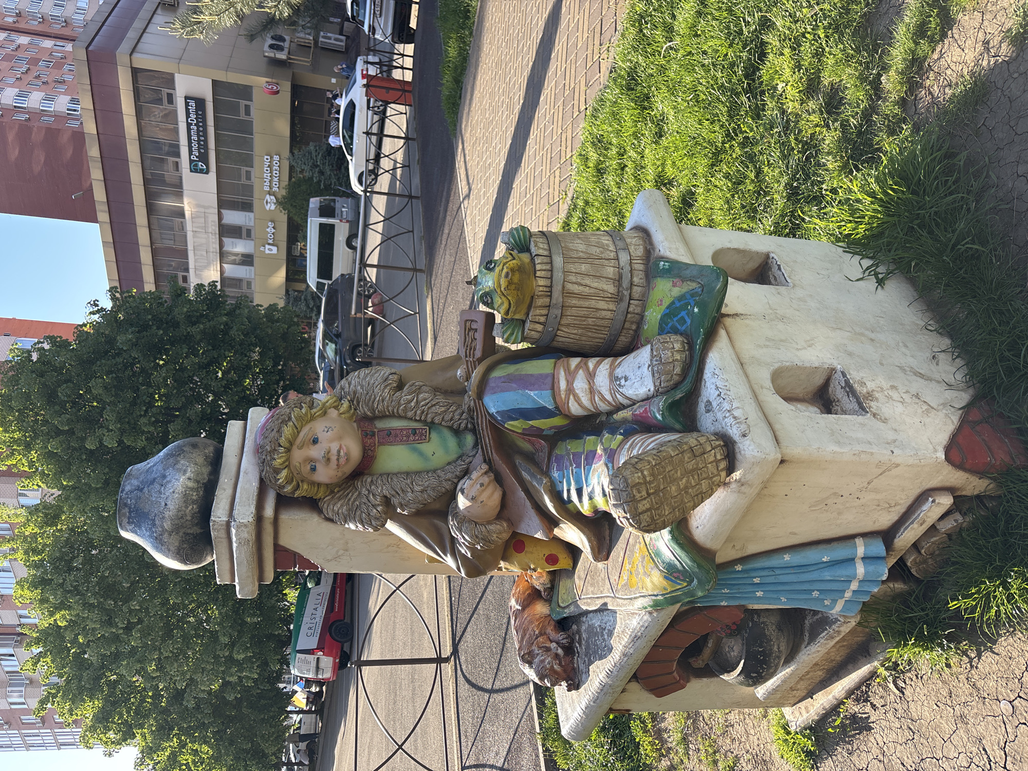 The image shows a colorful, folk-art-style sculpture of a sleeping figure with a barrel and other objects, situated on a sidewalk in Krasnodar, Russia. A building is visible behind the sculpture with a sign that reads "Panorama-Dental Diagnostic". The sidewalk is made of pavers and there is grass adjacent to the sculpture. The sculpture includes a dog. There are cars and pedestrians in the background.