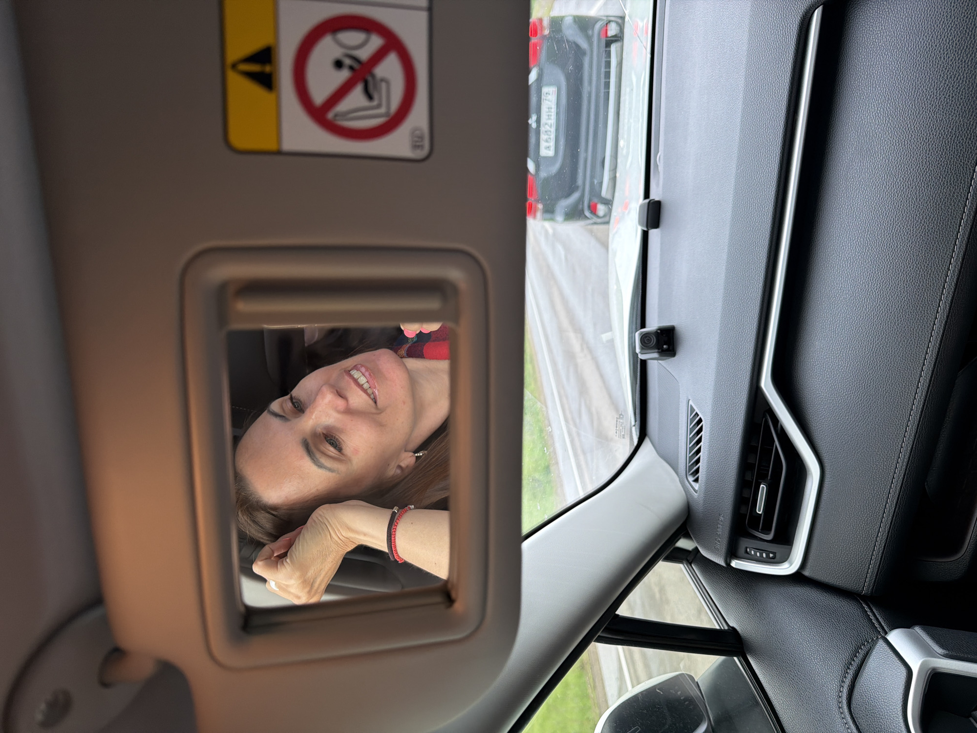 The image shows a reflection of a woman in a car's visor mirror. The woman has brown hair, is smiling, and is resting her left arm on her head. She is wearing a red and black plaid top and two bracelets on her left wrist. The car appears to be in motion on a paved road, as indicated by the blurred background which includes another car and a wet road surface. The interior of the car is visible, including the dashboard and an air vent. A yellow warning sticker with a red circle and a diagonal line over a silhouette of a person in a car seat is affixed to the visor. The image was taken in or near the city of Kirga, Russia.